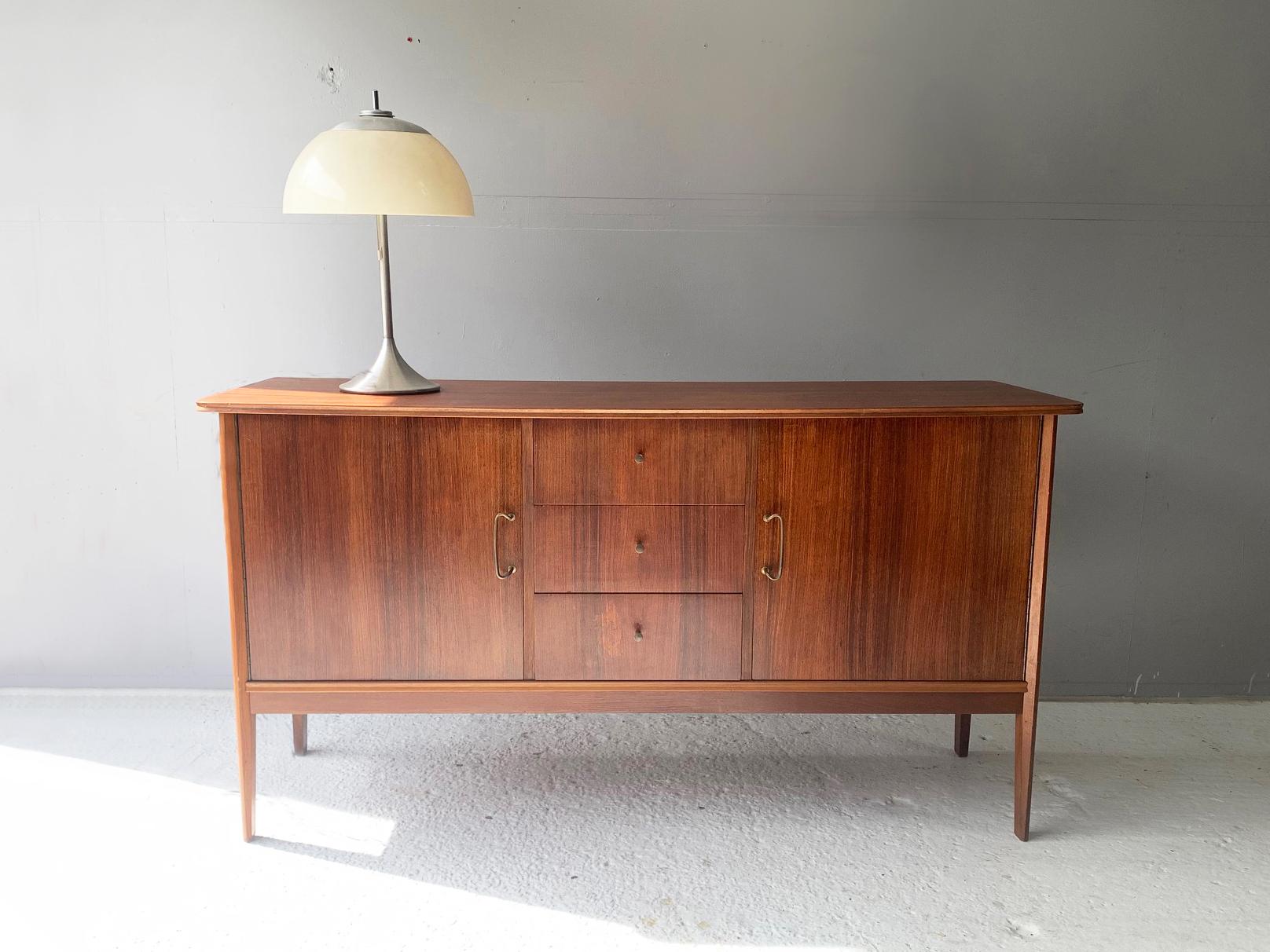 An elegant 1950s sideboard / buffet from British company Vanson, designed by Peter Hayward for Heal and Son. The sideboard is figured in walnut veneer onto a teak frame with three central drawers with brass trumpet handles.

Inside the top drawer