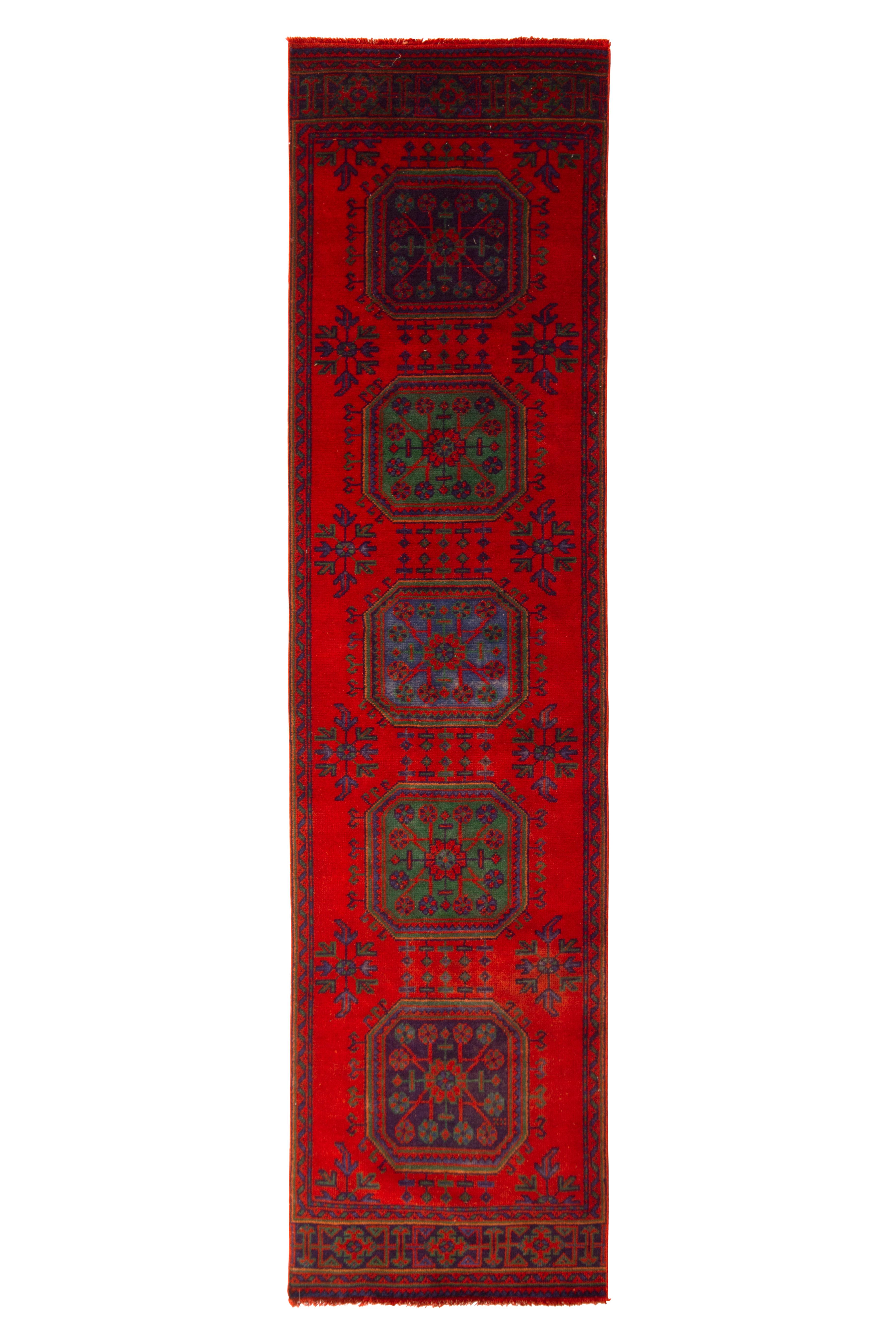 Hand knotted in wool from Turkey circa 1950-1960, this vintage mid-century Oushak runner employs a striking crimson red colorway, naturally juxtaposed with luminous green and blue hues bringing out a defined geometric floral emphasis in both the