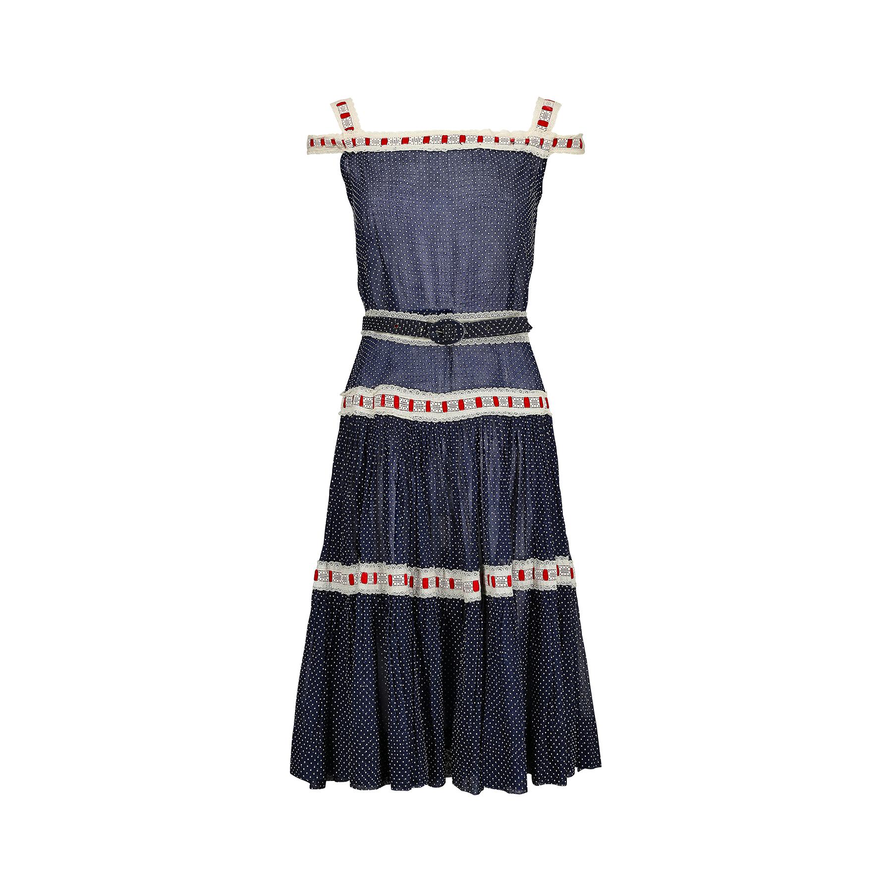 This is an unusual 1950s dress ensemble made from navy sheer cotton organza with a Swiss-dot print. It is trimmed with cream eyelet lace and red velvet ribbon work that is reminiscent of embellishment from the 1920s and 30s. This trim makes up the
