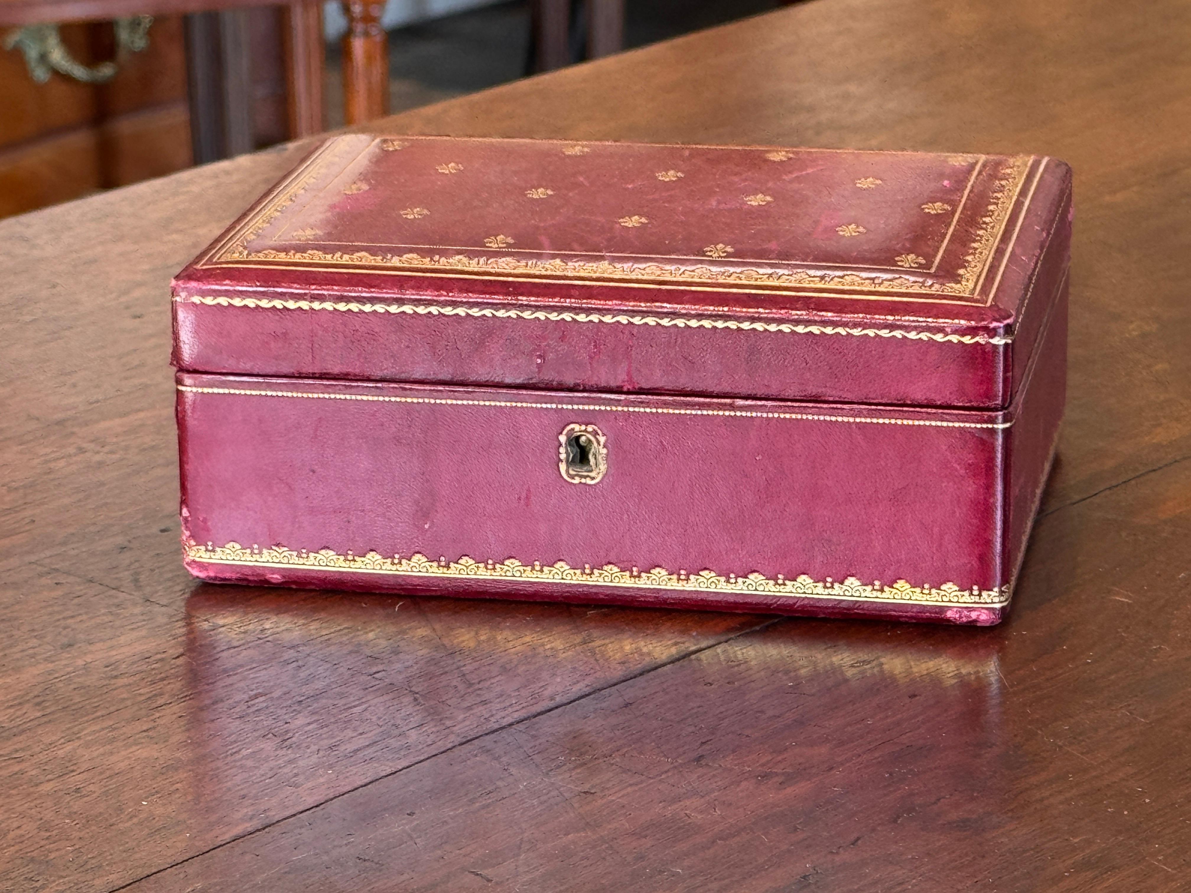 Boîte à bijoux en cuir rouge sang avec détails dorés et intérieur en velours rouge. Le plateau qui se soulève révèle un compartiment plus profond, tous deux revêtus d'un velours doux. Il conserve le trou de serrure d'origine et la délicate