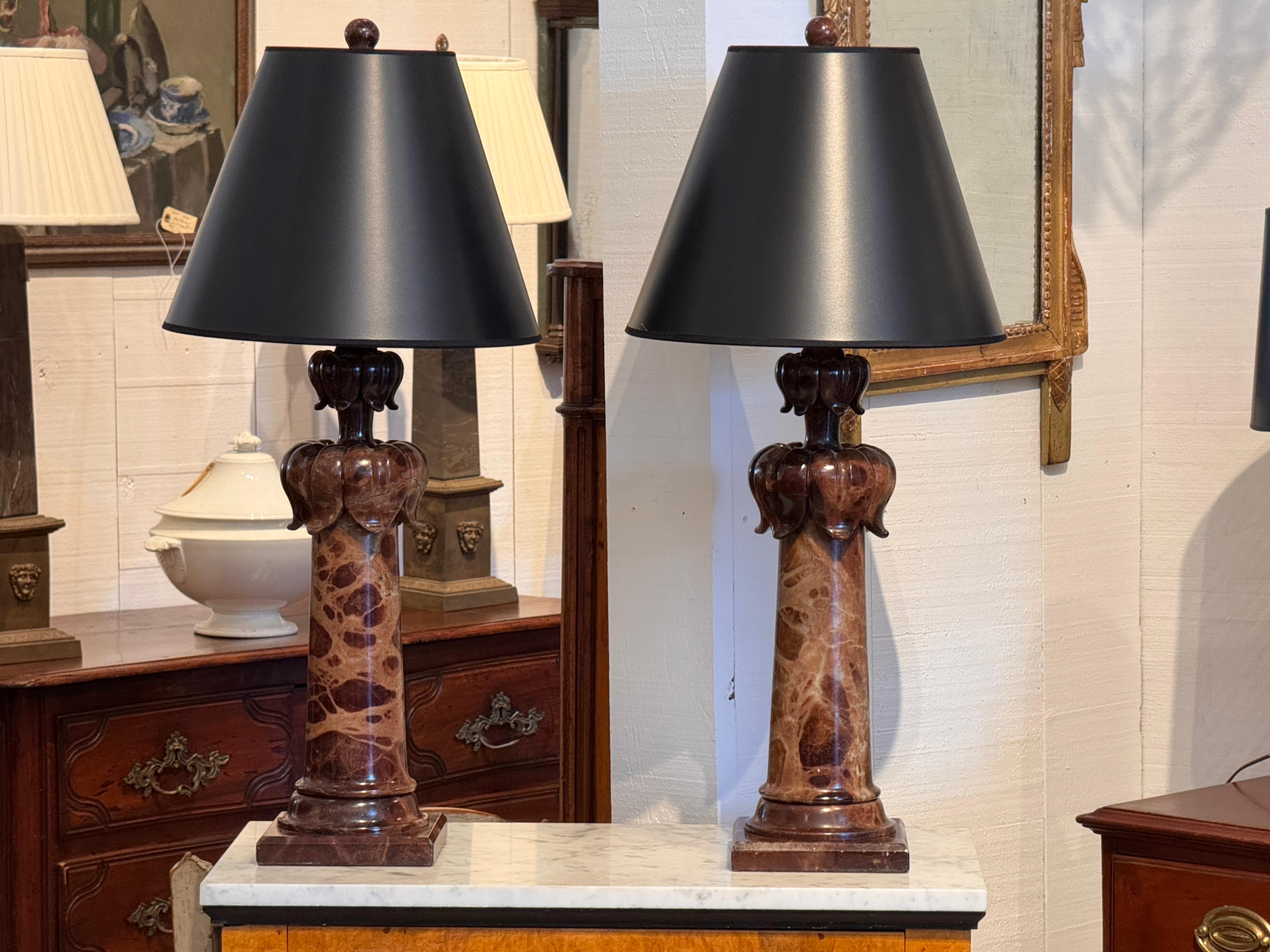 Coppia di lampade a colonna in alabastro dai toni caldi con basi quadrate a gradini. Ogni lampada presenta un capitello intagliato con dettagli lobati arrotondati sopra il fusto affusolato, con venature naturali e variazioni di colore tipiche