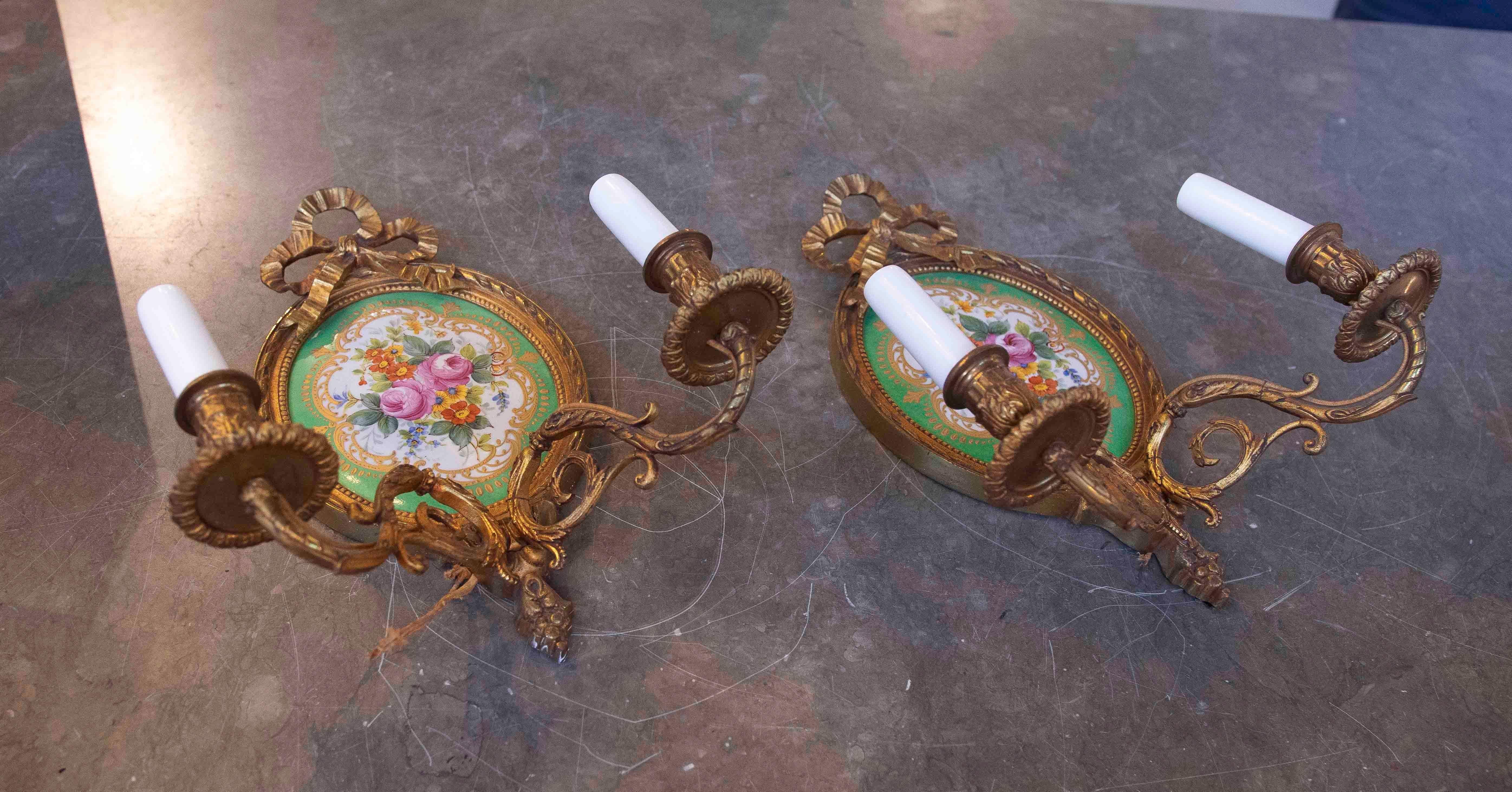 Coppia di appliques in bronzo dorato e porcellana, ornate da un medaglione centrale decorato con motivi floreali dipinti a mano su sfondo verde. I bordi in bronzo dorato e il dettaglio del fiocco in alto incorniciano il design, aggiungendo un tocco