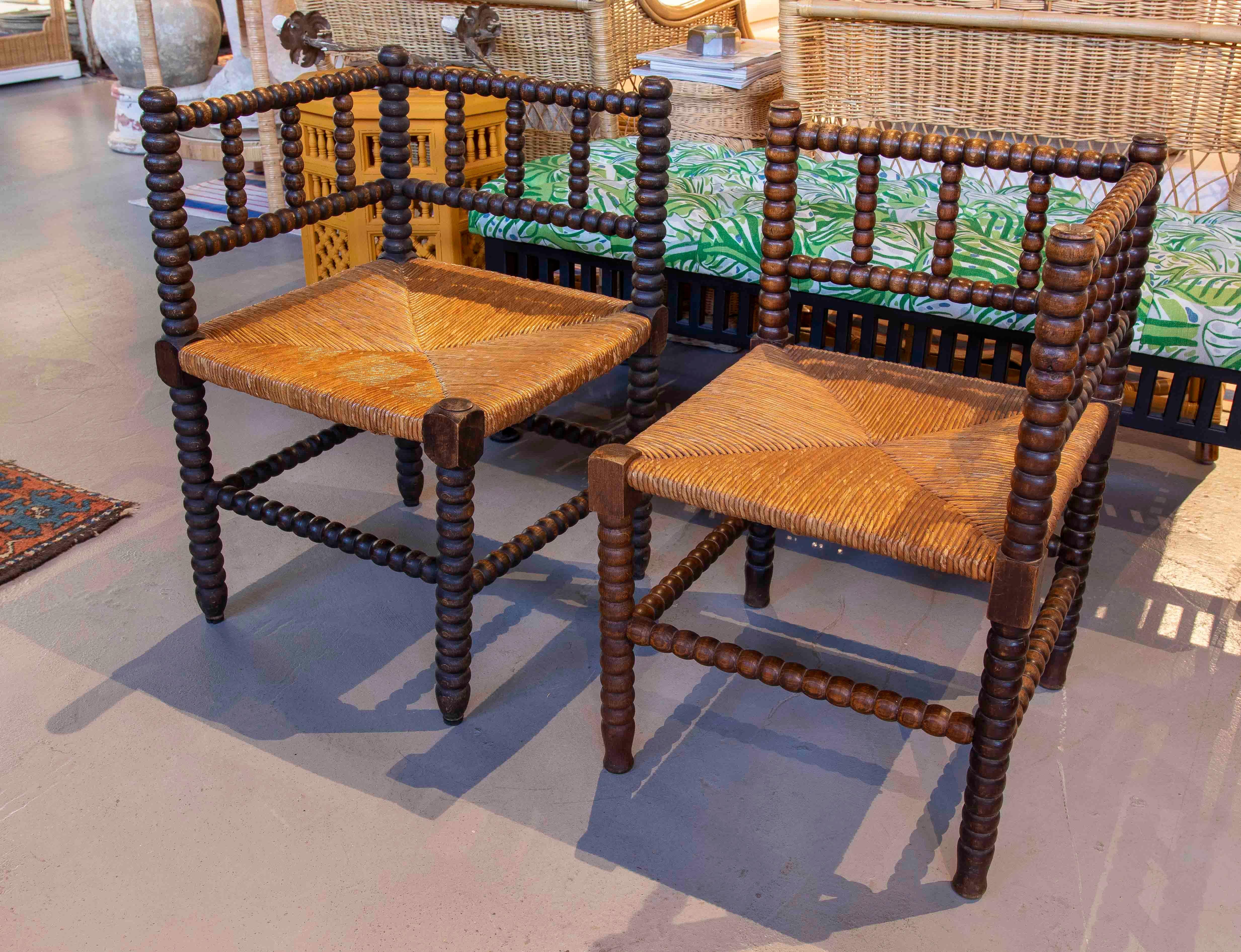 Pareja de sillas esquineras con armazón de madera torneada en forma de huso y respaldo enmarcado. Los asientos son de mimbre tradicional tejido a mano, que ofrece un contraste cálido y natural con el acabado oscuro de la madera. Su diseño simétrico