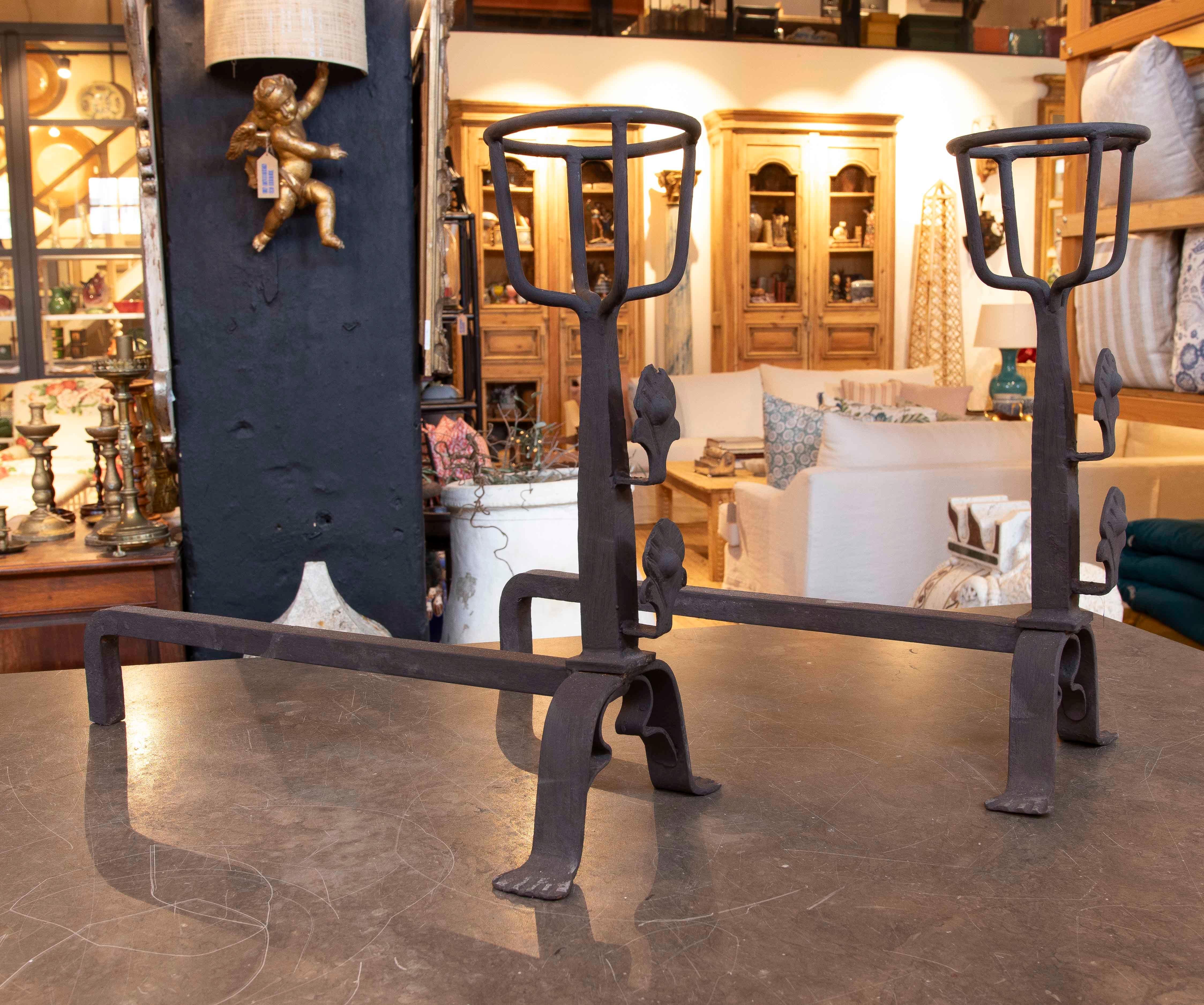 Ein Paar schmiedeeiserne Andirons mit ornamentalen Details in Form von Blättern, die zur Aufnahme von Brennholz in traditionellen Kaminen dienen. Ihre robuste Struktur und ihr dekoratives Design machen sie zu funktionalen und ästhetischen Stücken,