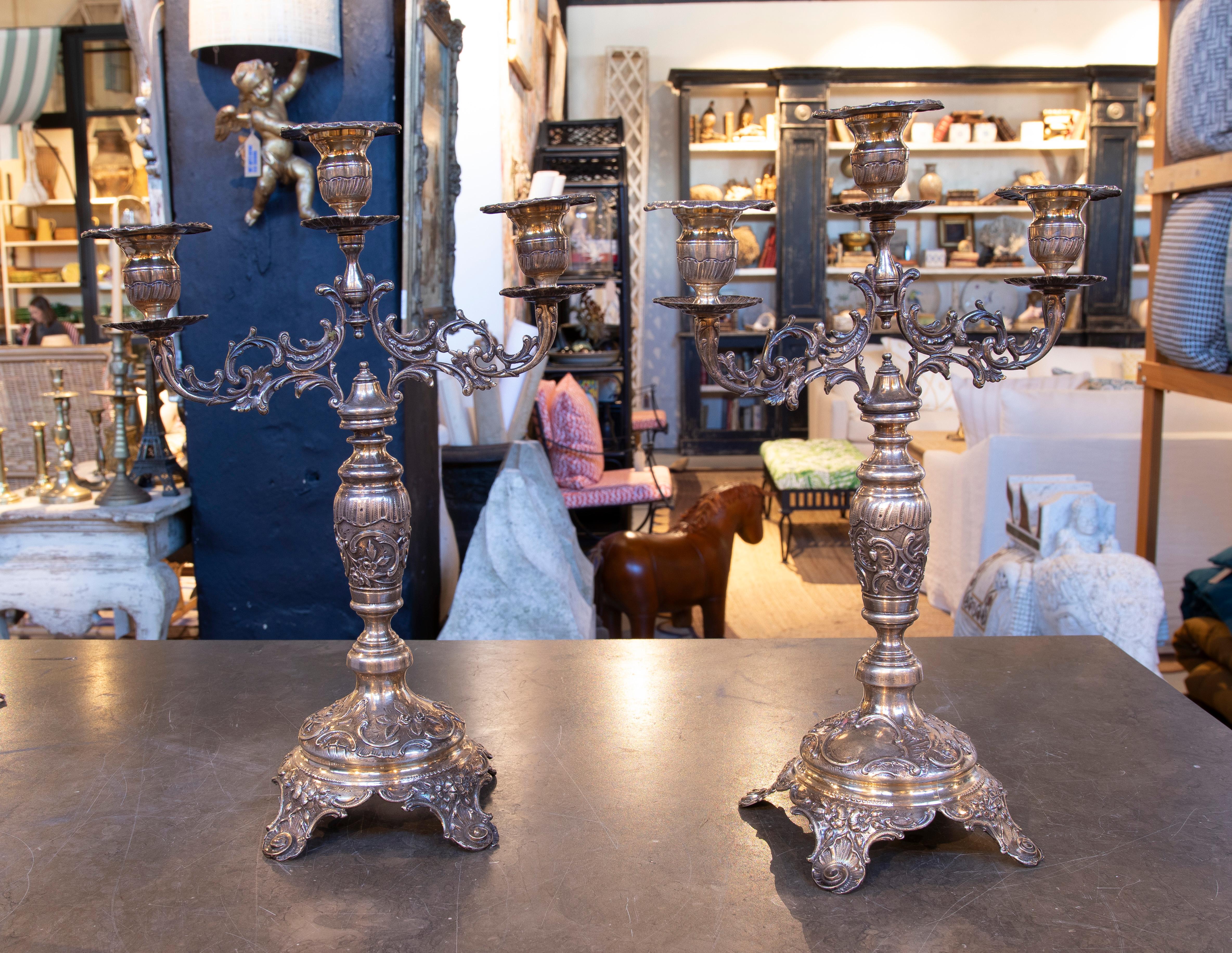 Una squisita coppia di candelabri d'argento con un design barocco ornato. Ogni candelabro presenta cinque bracci riccamente decorati con dettagli floreali e curve stilizzate, sostenuti da una robusta base scolpita con motivi classici. La patina