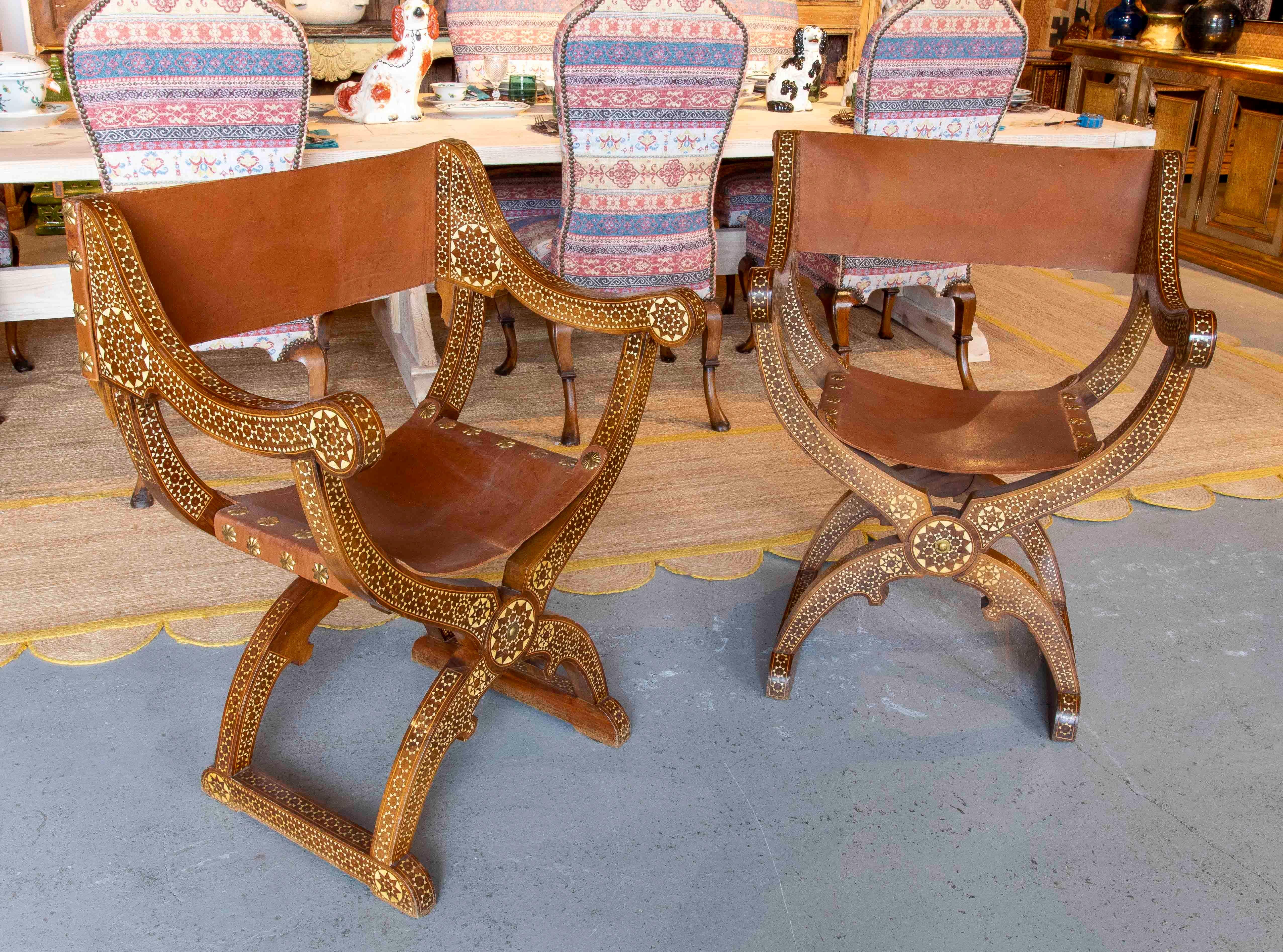 Une élégante paire de chaises de style Curule, fabriquées en bois sculpté avec des incrustations d'os et de nacre dans de délicats motifs géométriques. L'assise et le dossier sont en cuir naturel, tendu sur le cadre, ce qui apporte confort et