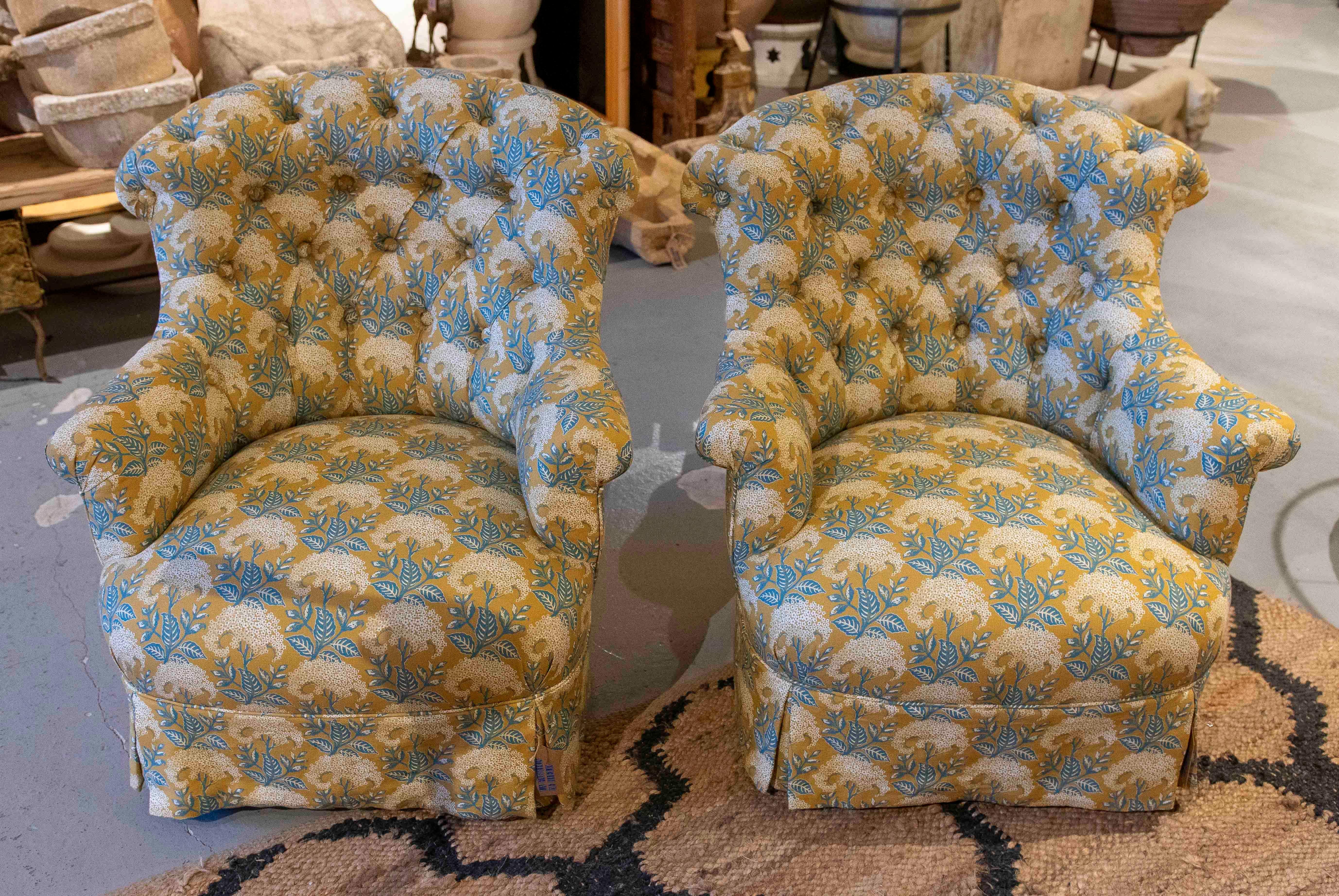 Un encantador par de sillones vintage tapizados con un elegante estampado floral en tonos azules, dorados y marfil. Sus respaldos acolchados con botones, brazos suavemente curvados y bases con faldón aportan un aire clásico y confortable, perfecto