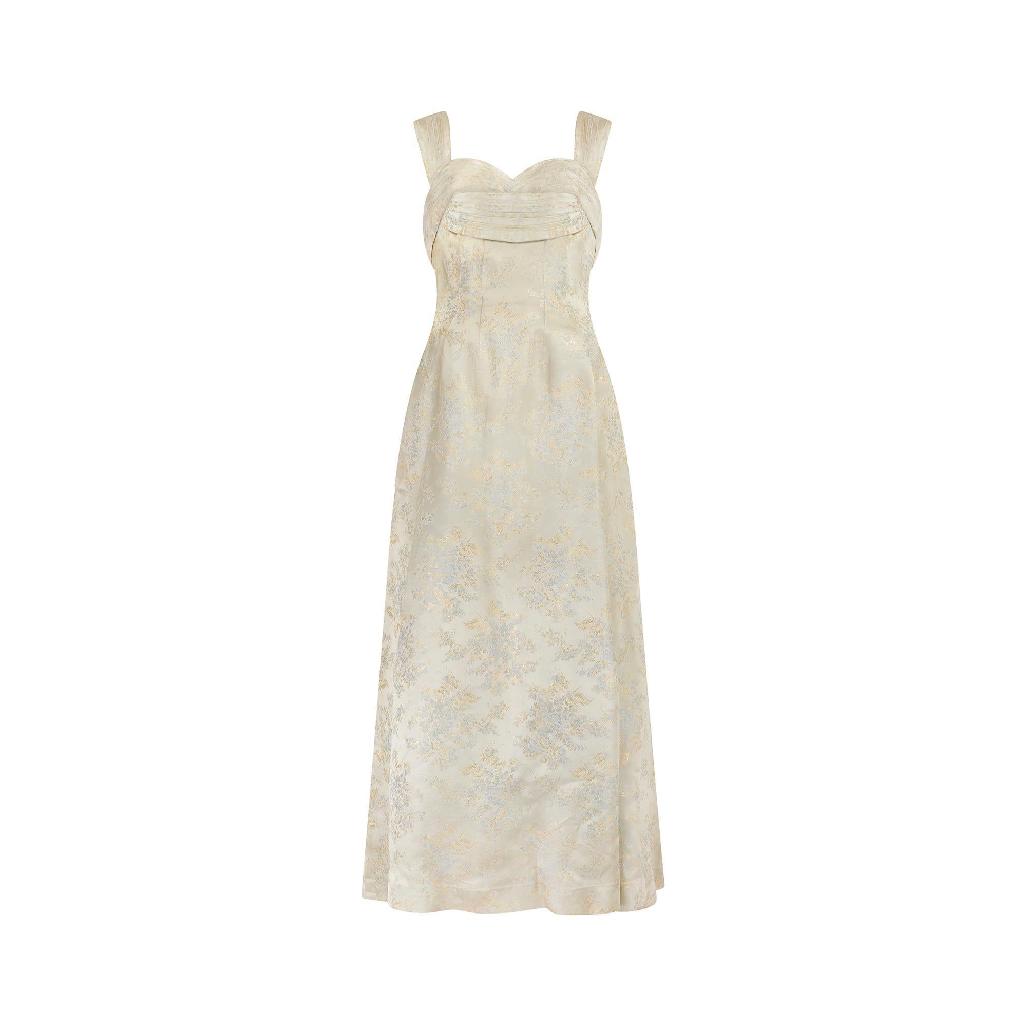 This British couture made jacquard ball gown dates to the mid 1950s and has been finished with hand-sewn details. The shimmering champagne hued brocade is detailed with gold ferns and blue-grey flowers. The wide straps and bust are intricately