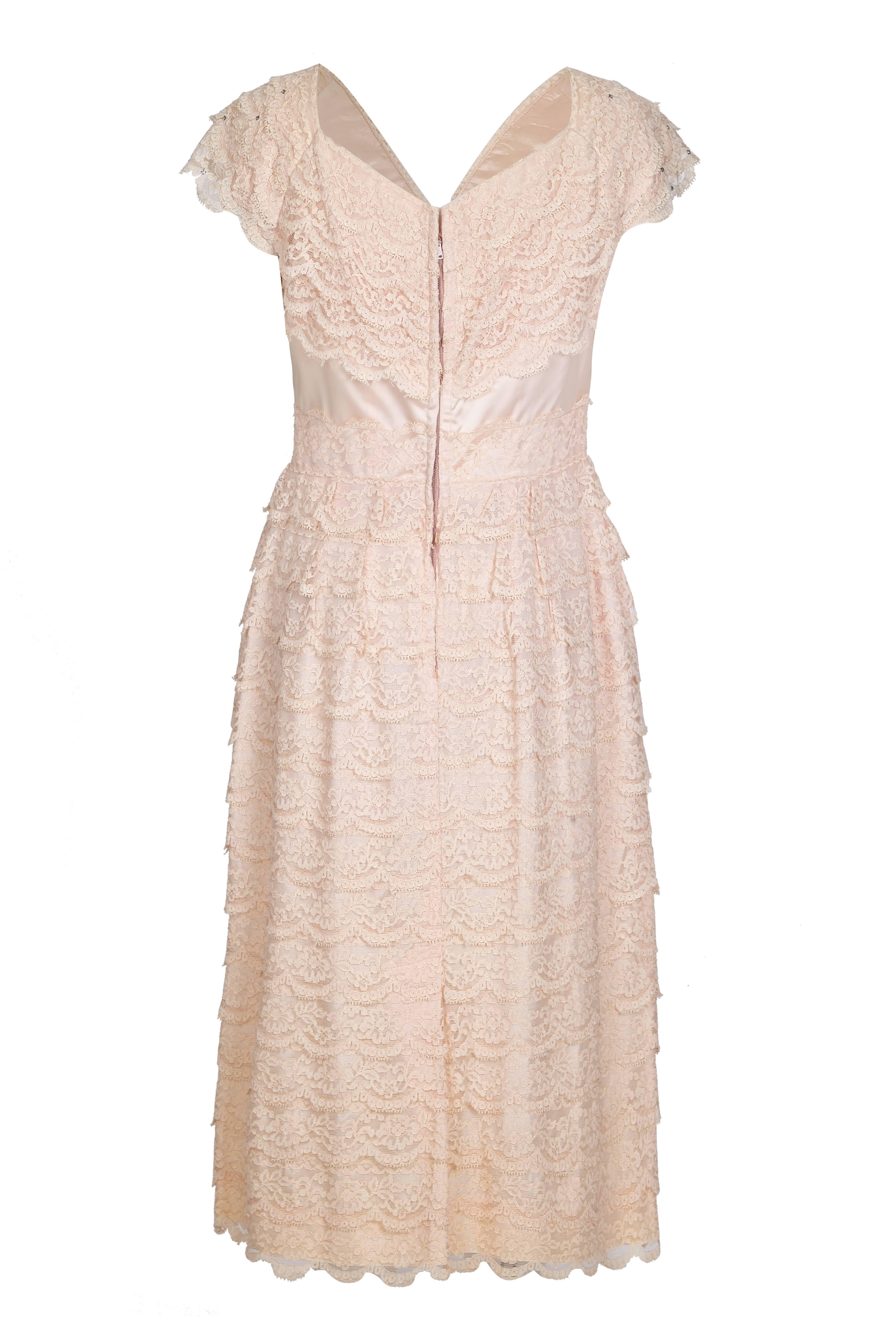 This charming vintage 1950s pale pink tiered lace dress is gamine, playful, and in lovely vintage condition. Designed to flatter an hourglass figure, the dress has a simple line fitting snugly at the waist and with generous give in the hips and