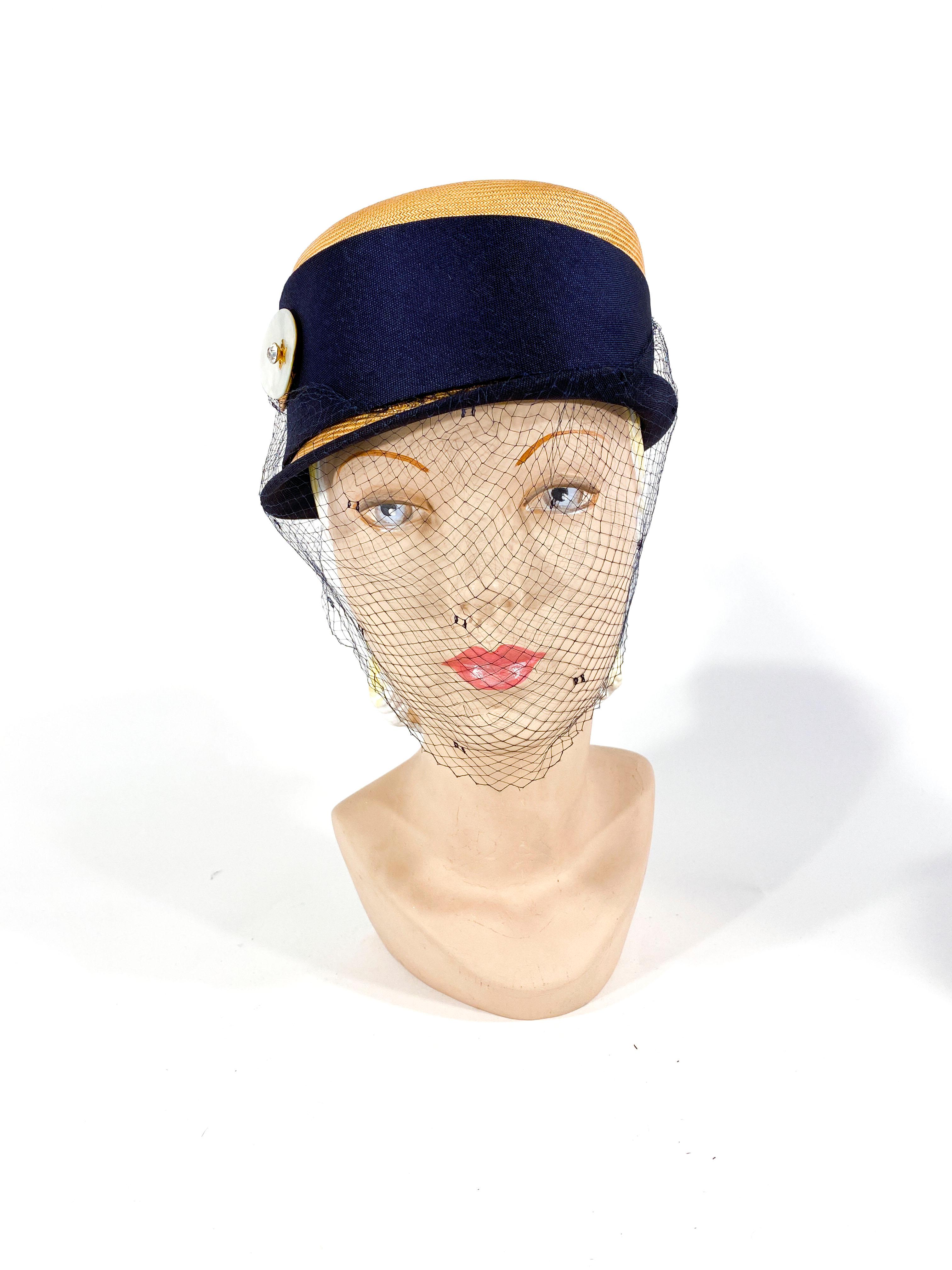 1950s natural panama woven modified pill box hat with a side brim, navy edging, a wide asymmetrical band. The band is sculpted with a hand cut abalone and jeweled button. The full-face veil is very dark navy blue and has a tie back function to