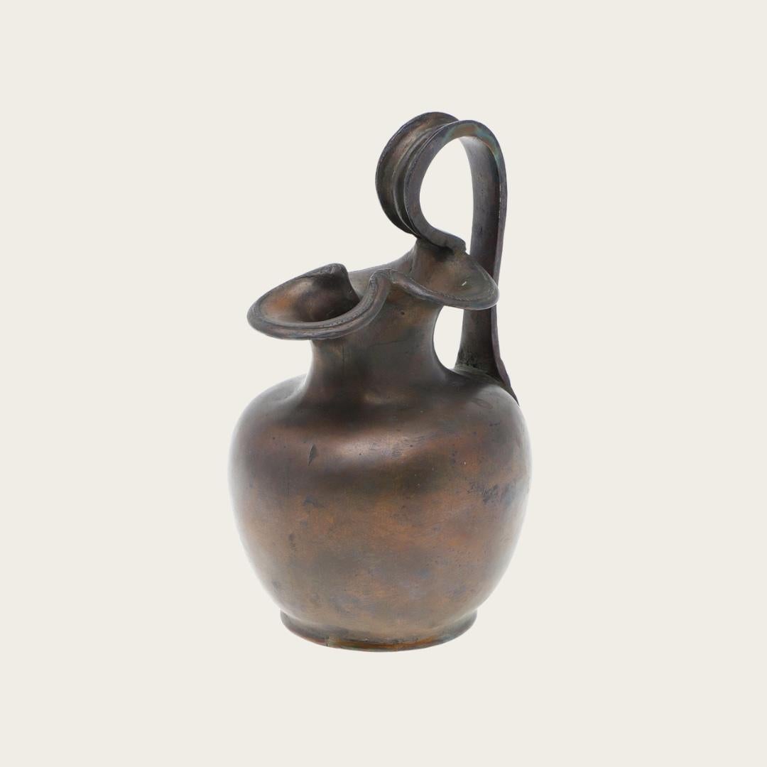This 1950s patinated bronze vessel is designed in the style of an ancient Greek oenochoe. Its decorative handle features a detailed angelic figure, adding a unique and artistic element. The aged bronze patina enhances its vintage appeal.