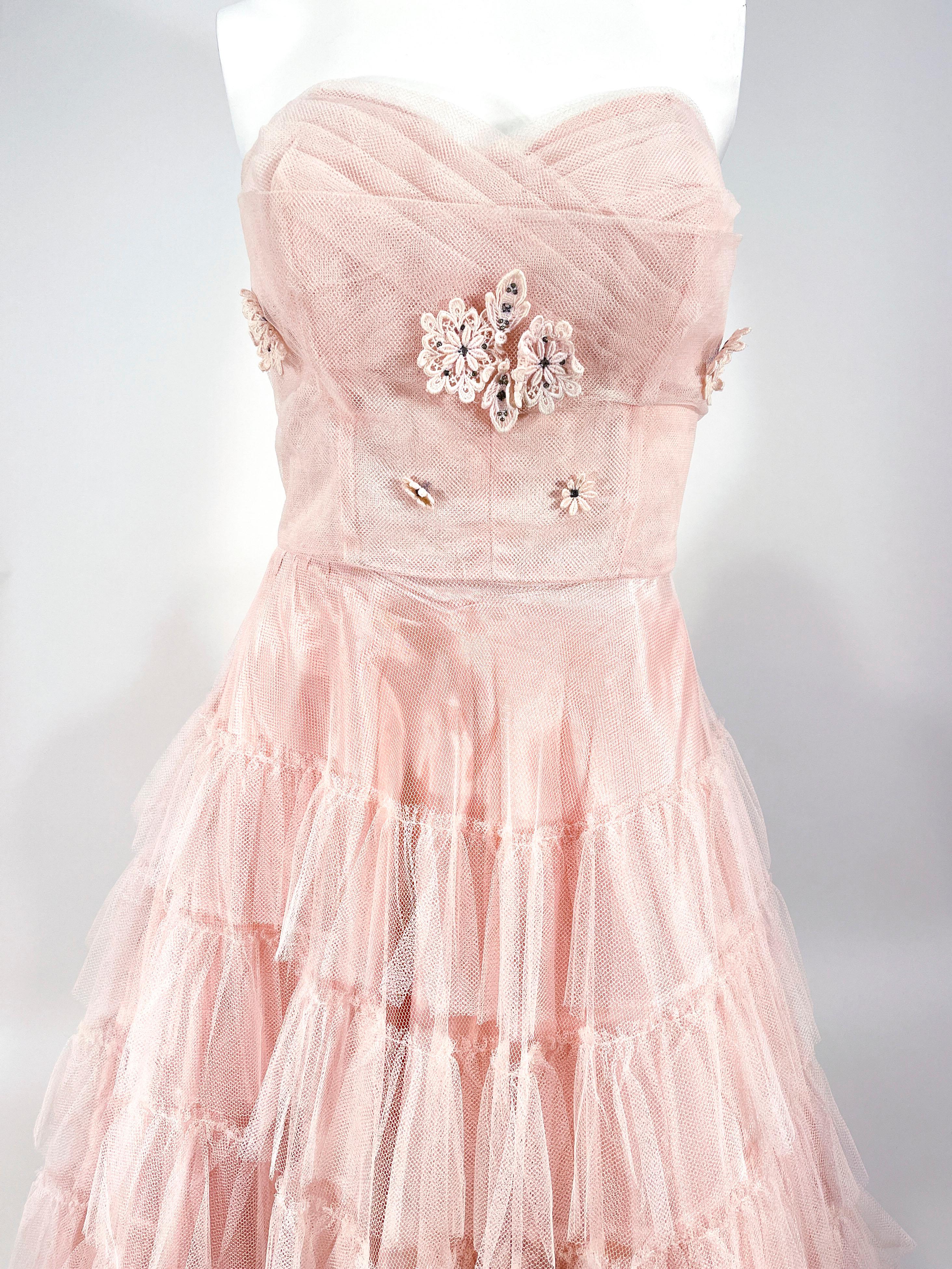 1950s pink tulle party dress featuring a sleeveless bodice decorated with pleated tule and pink appliqué with added rhinestones. The full skirt has multiple layers of ruffled tulle that go down to a tea length. This dress is fully lined in taffeta