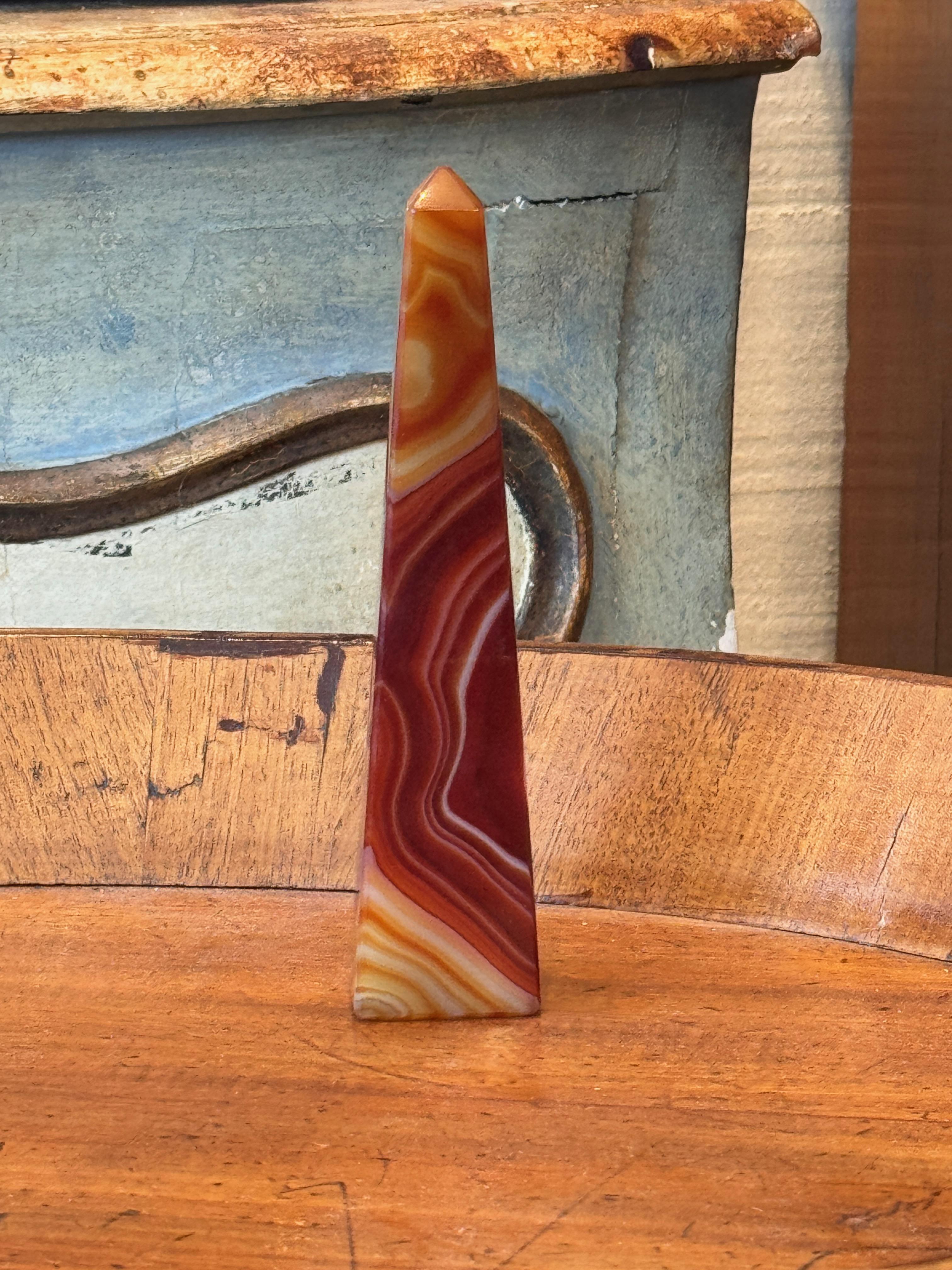 Pequeño obelisco esbelto tallado en ágata bandeada, que muestra estrías naturales en tonos cálidos de rojo, naranja y crema. La superficie está finamente pulida para resaltar las capas vivas y los patrones orgánicos en toda la superficie