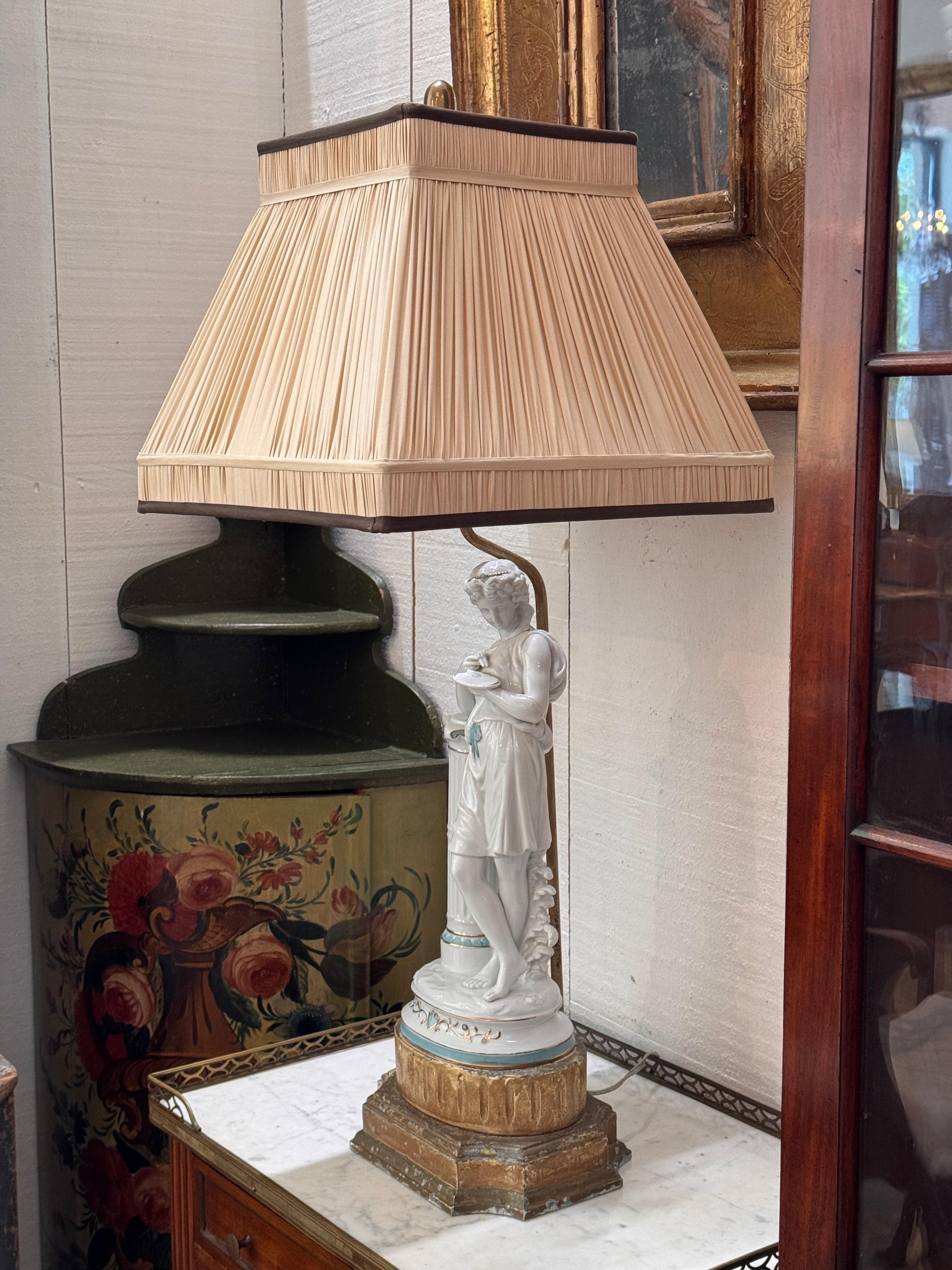 Lampe de table figurative en porcelaine représentant une jeune fille drapée de façon classique, appuyée sur une colonne cannelée. La figure est rehaussée de détails sarcelle pâle et dorés et repose sur une base en bois doré à gradins. Surmontée d'un