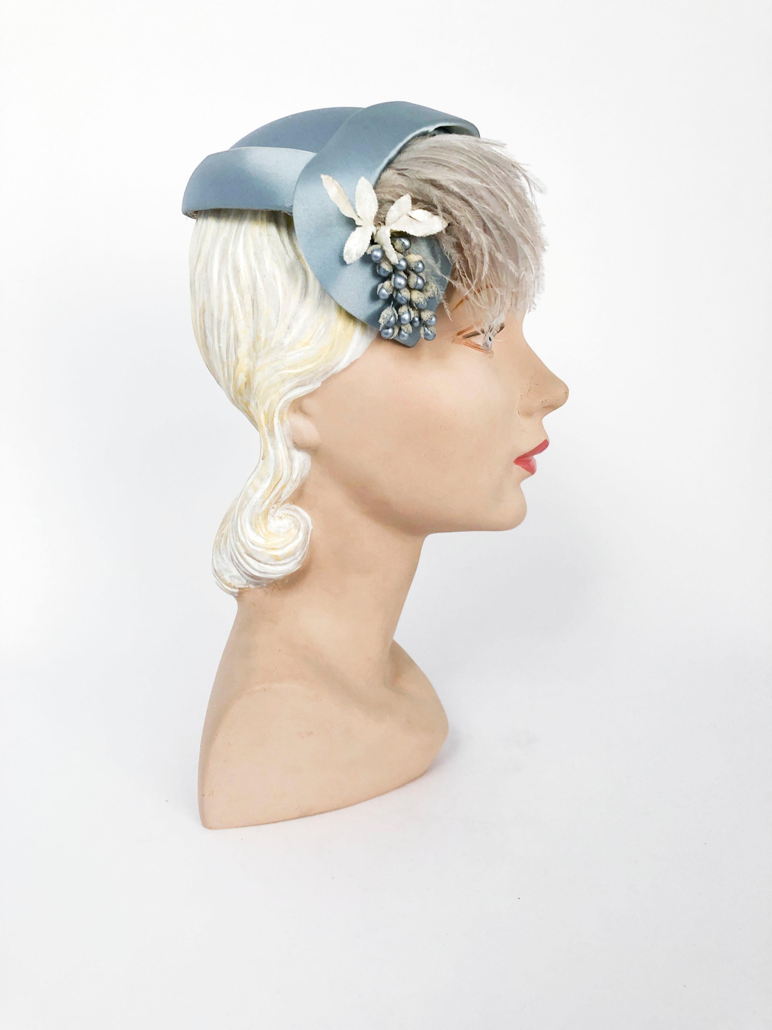 1950s Silk satin hat in powder blue with an asymmetrical shaping, velvet leaves, fruit accent, and feathered wisp. Combs on the band to further secure the hat to the head