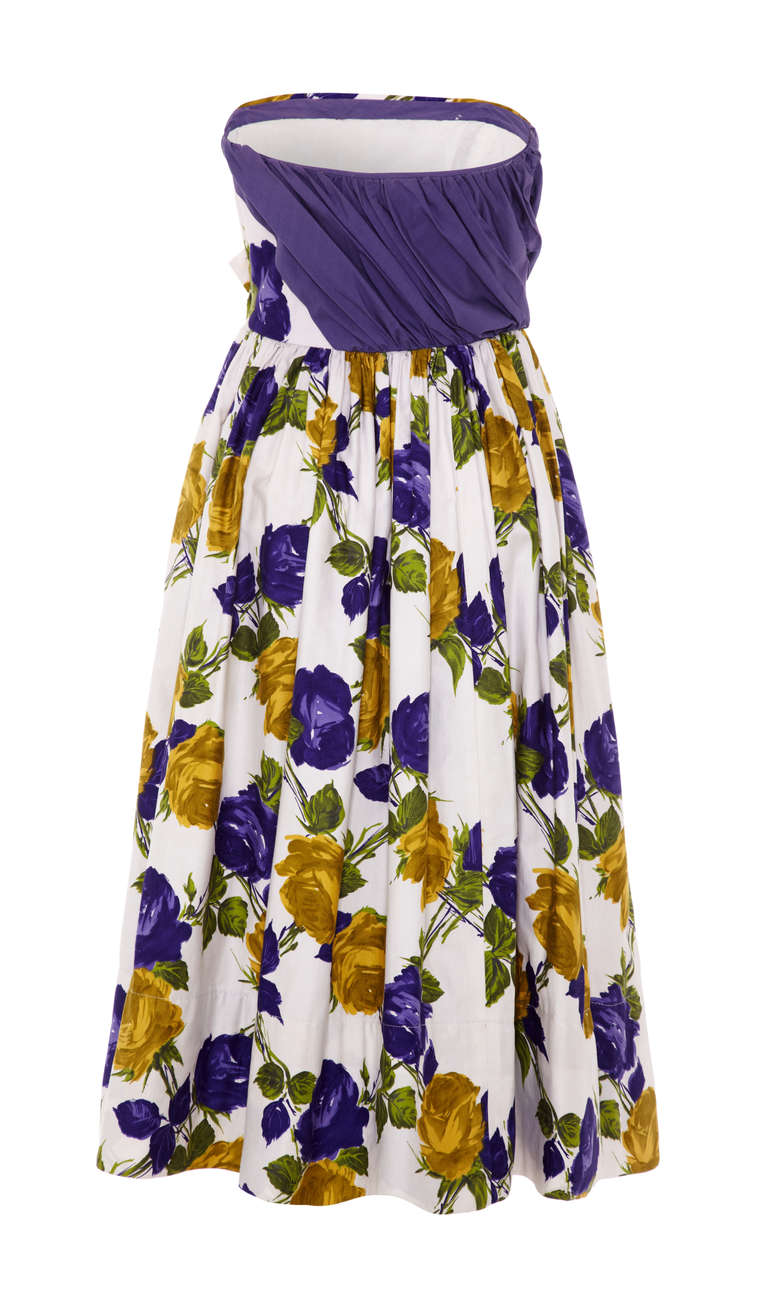 A lovely and summery original 1950s white cotton dress featuring a large rose print in yellow, purple and green. The strapless bodice is asymmetrical and has a pretty gathered purple section wrapped around one side and a bow to the front in matching