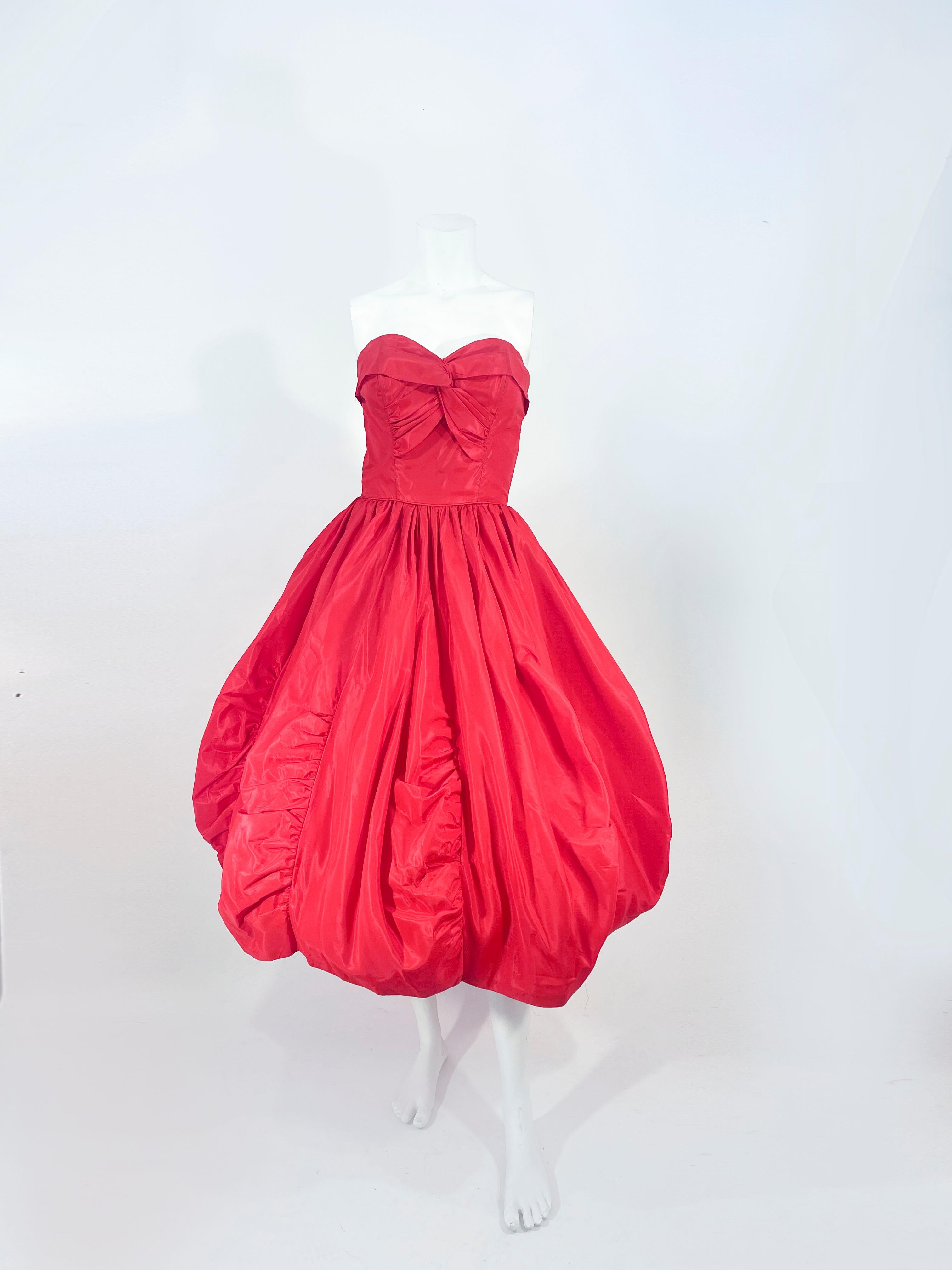 Rotes Partykleid aus Taft der 1950er Jahre mit gerüschtem und getäfeltem Bubble-Saum. Das Mieder ist mit Falten und Raffungen verziert, hat innenliegende Stäbchen für mehr Struktur und eine Taillenschürze. 

Der Rücken hat einen Metallreißverschluss
