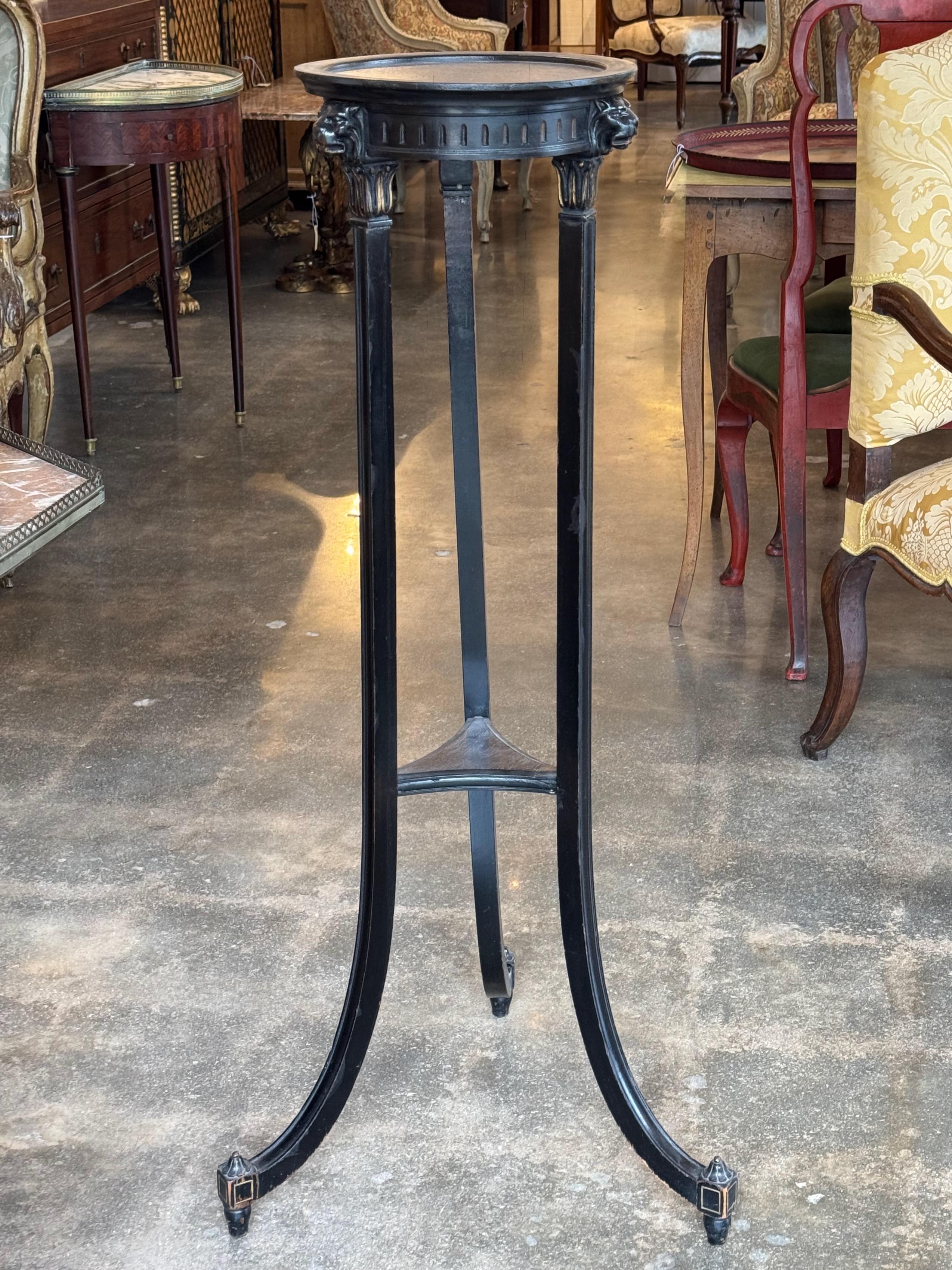 Alto ed elegante piedistallo dipinto di nero in stile Regency, caratterizzato da tre sottili gambe affusolate unite da una base a barella. Il piano circolare è sostenuto da capitelli intagliati con abbellimenti a maschera di leone, che aggiungono un