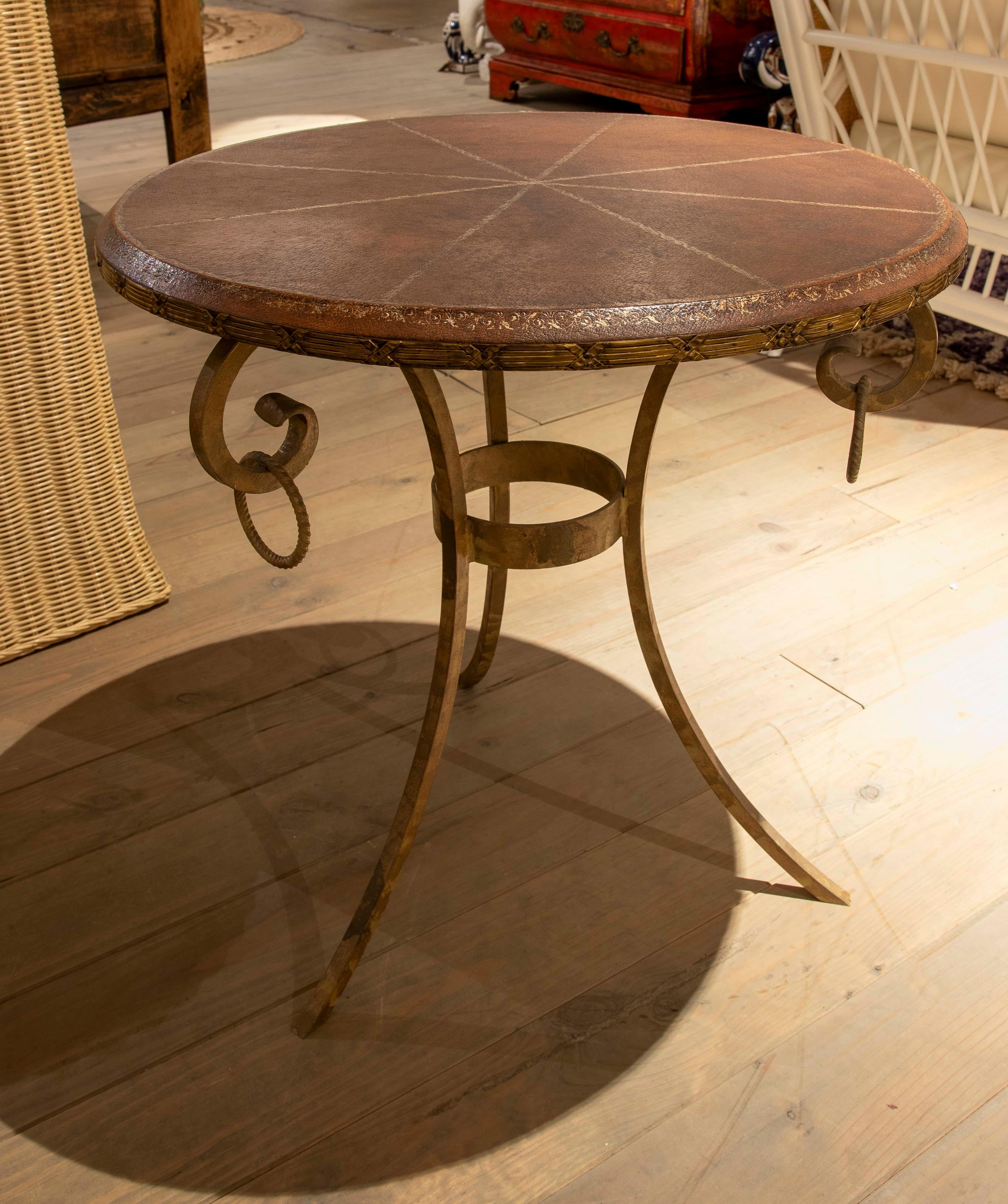 Elegante tavolino artigianale con piano rotondo rivestito in pelle marrone goffrata, decorato con sottili motivi geometrici. La struttura in ferro battuto è caratterizzata da sottili gambe ricurve e da dettagli a spirale, che garantiscono stabilità
