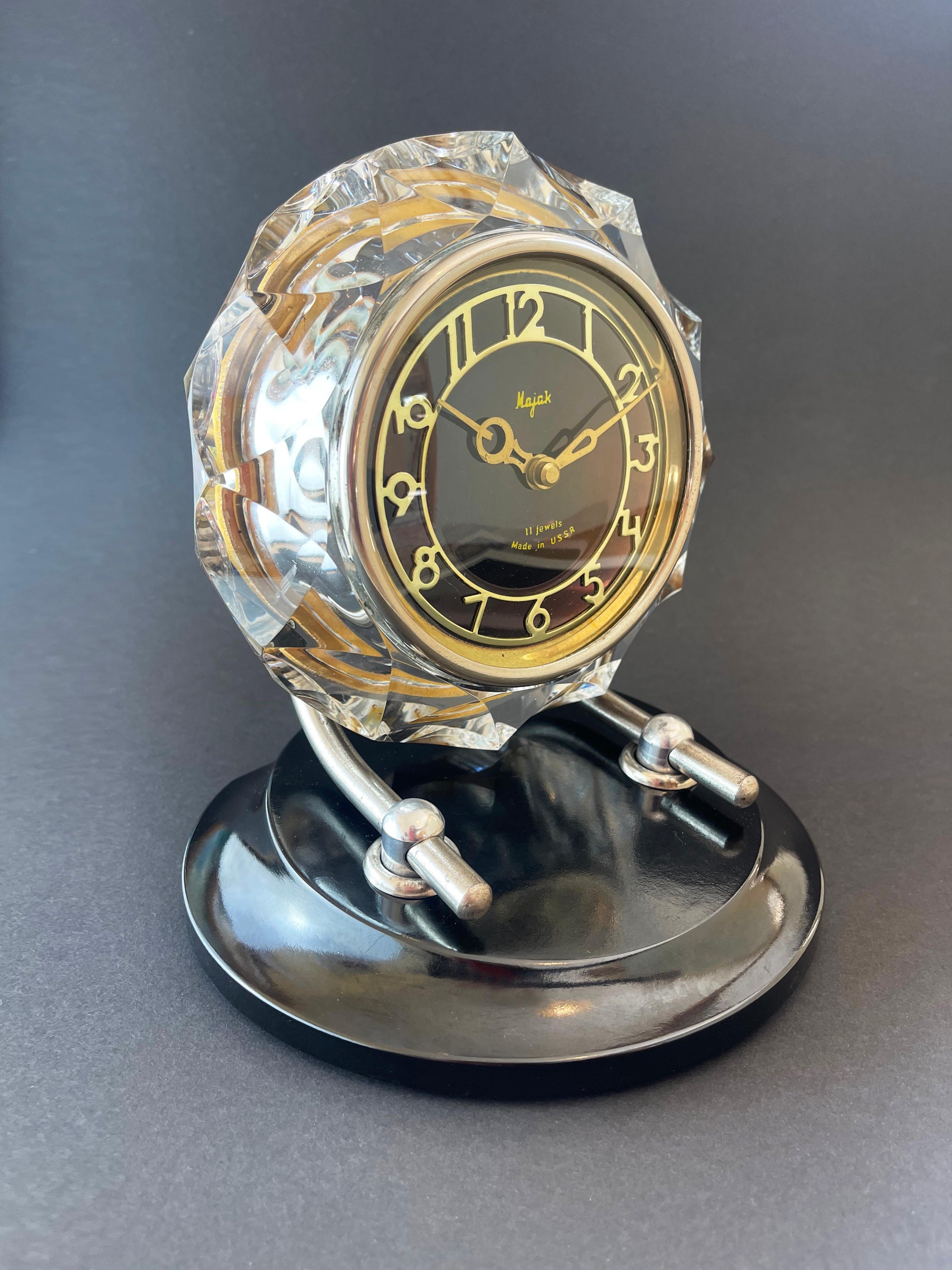 An impressive art deco style has been implemented in this mantel or table clock:
the chunky cut crystal glass encompassing the golden shimmering brass dial on black ground.
The bakelite base holds the clock itself on two adjustable silver metal