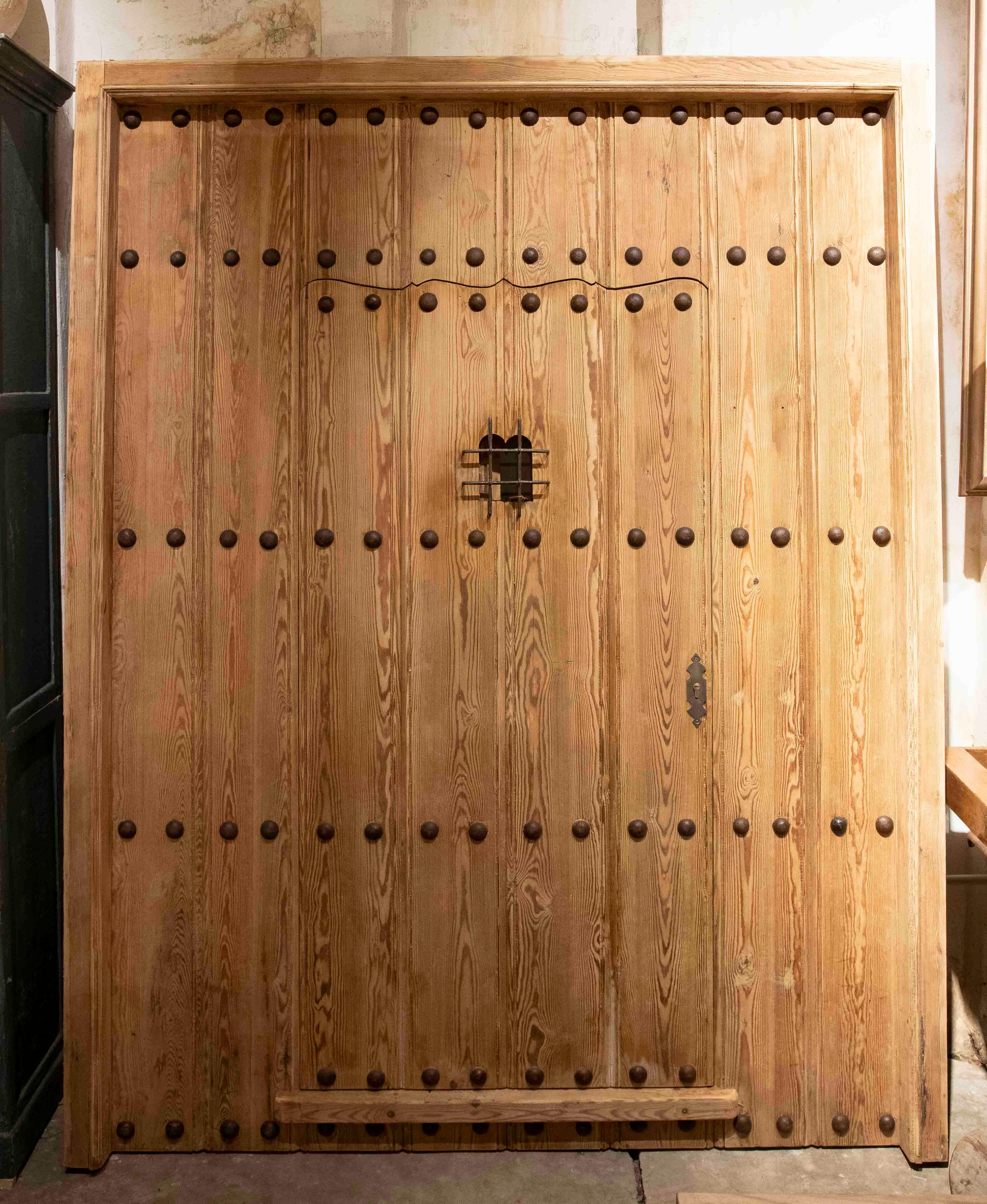 Una imponente verja rústica española de madera maciza, compuesta de tablones verticales ensamblados con precisión y decorados con clavos forjados de gran tamaño dispuestos simétricamente. Su diseño evoca la arquitectura tradicional de casas rurales,