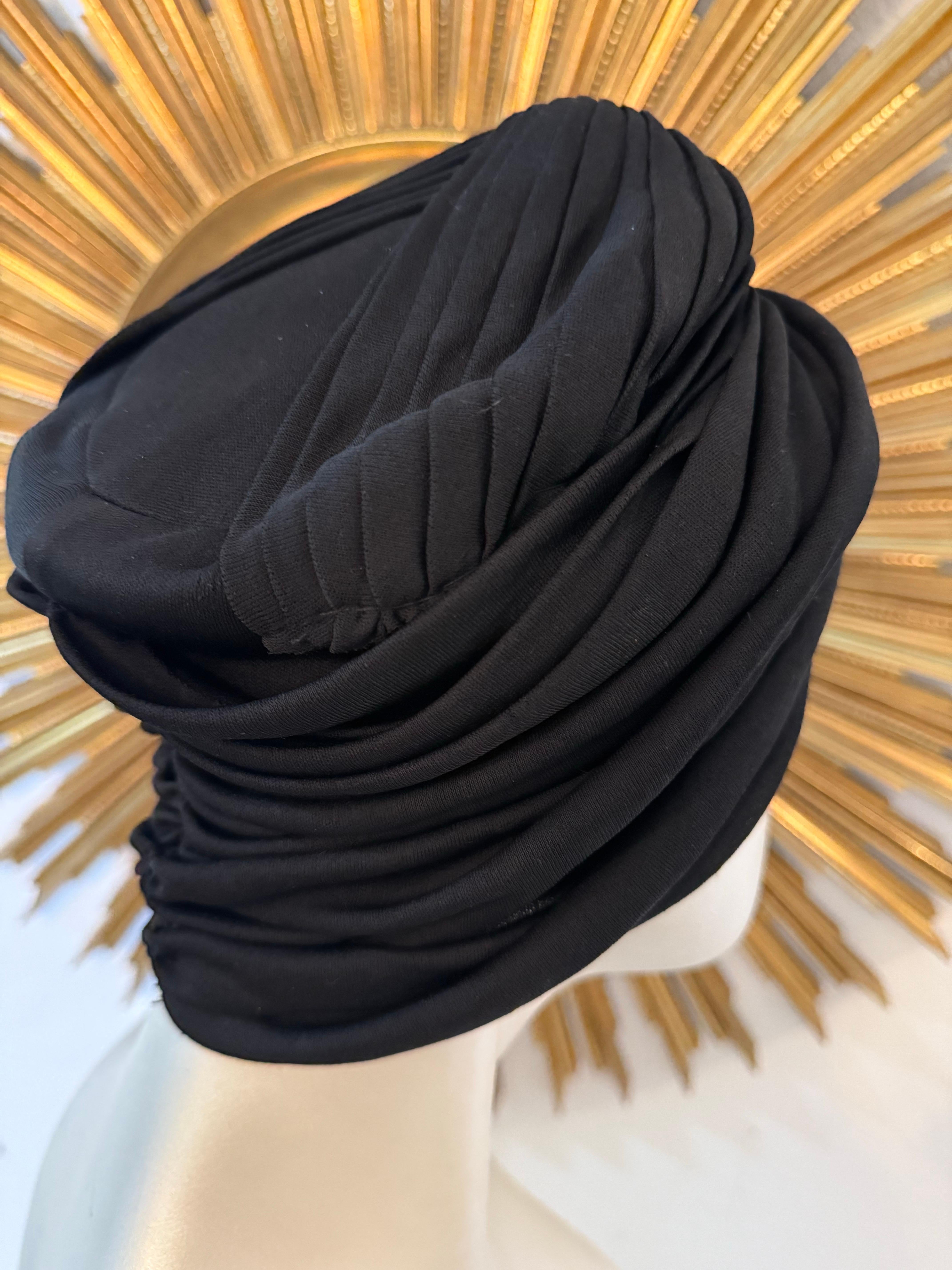 1950s Saks Fifth Avenue Black Silk Matte Jersey Draped and Knotted Turban: Features a structured, lined and pleated crown top. Sides are unstructured to fit over the ears. Gathered at center back. One size fits all. 