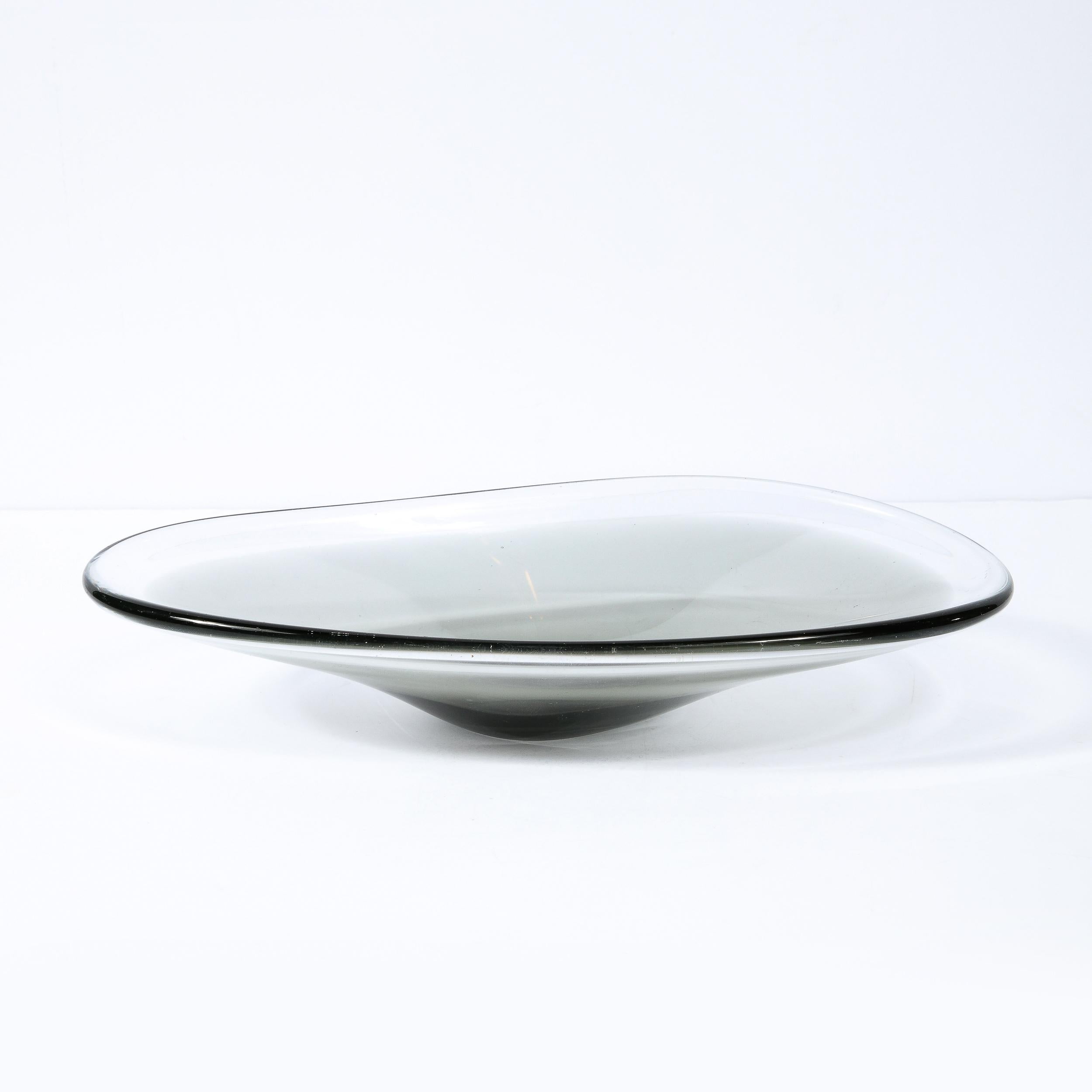 This elegant Scandinavian Mid-Century Modern art glass bowl was realized by the esteemed maker Holmegaard in Sweden circa 1950. It features an elliptical silhouette with raised sides and a concave center in a sultry smoked semi translucent art