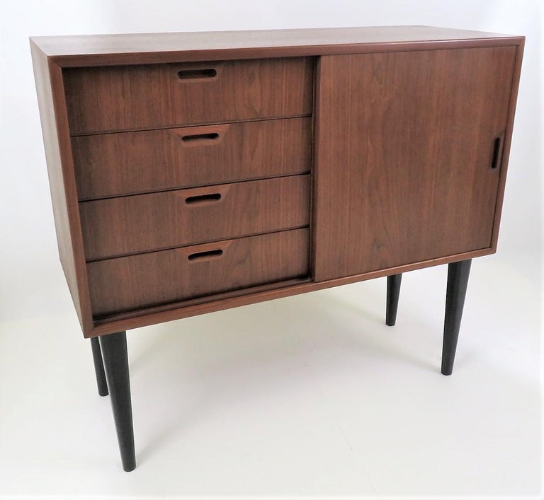 Very nice smaller scale walnut credenza, bedside cabinet or storage cabinet with 4 drawers and opposite, an 18 inch wide adjustable shelf behind the sliding door. Black ebonized tapered legs complement the design of this fine Danish made cabinet