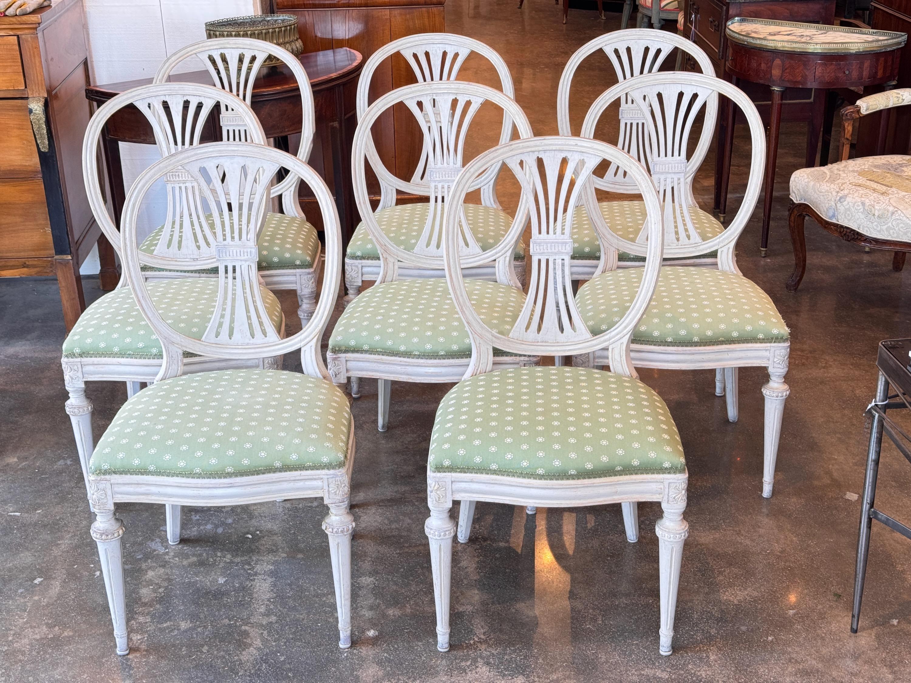 Ensemble de huit chaises de salle à manger peintes avec dossiers en forme de lyre et rosettes sculptées aux angles. La finition blanchie à la chaux est associée à des sièges rembourrés verts avec un motif floral à petite échelle. Les pieds fuselés