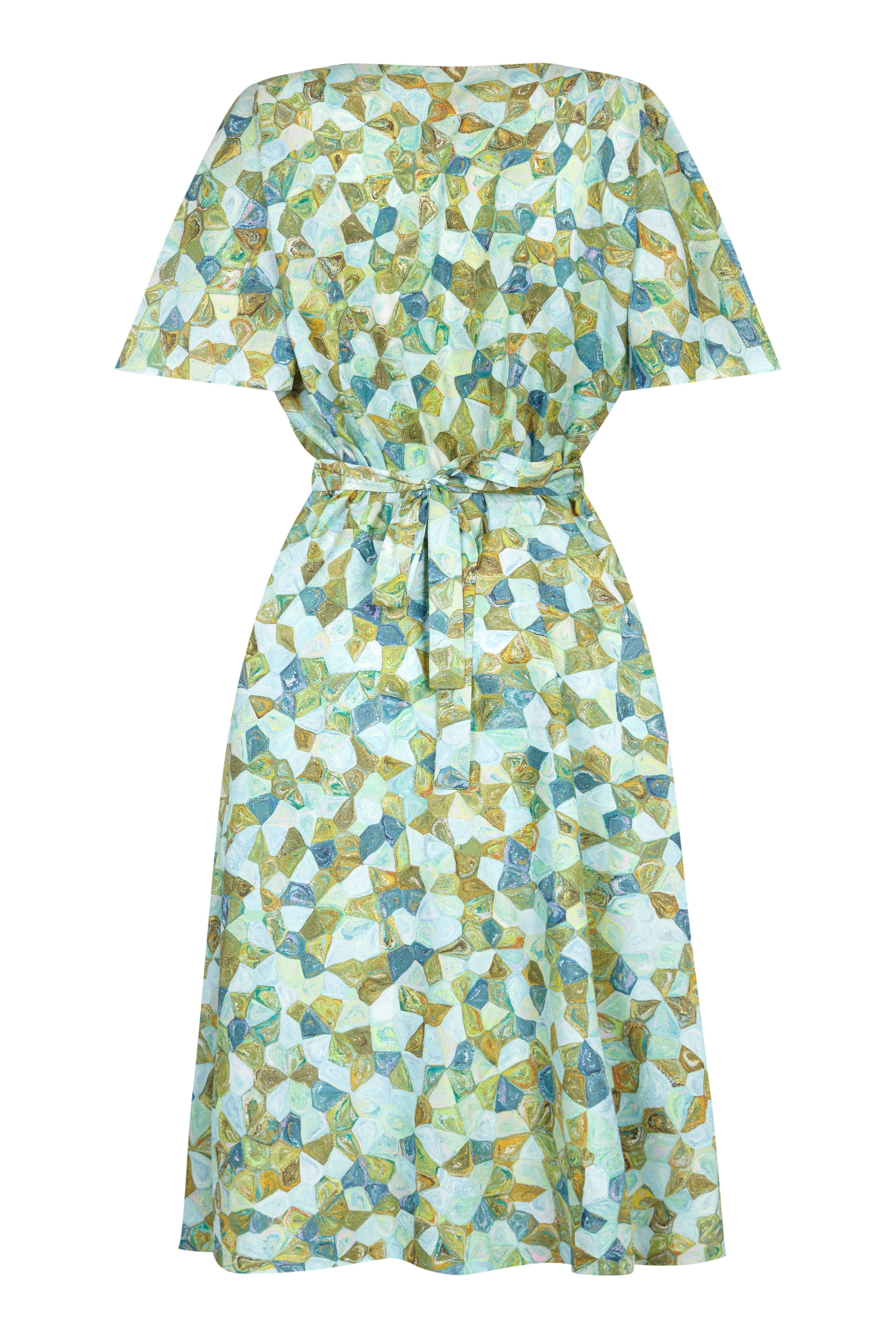 This pretty 1950s silk green patterned dress features an unusual kaleidoscope design in a variety of attractive shades of green and pale blue. The dress is deeply feminine with a full, ankle length skirt, and flared shirt sleeves that create soft