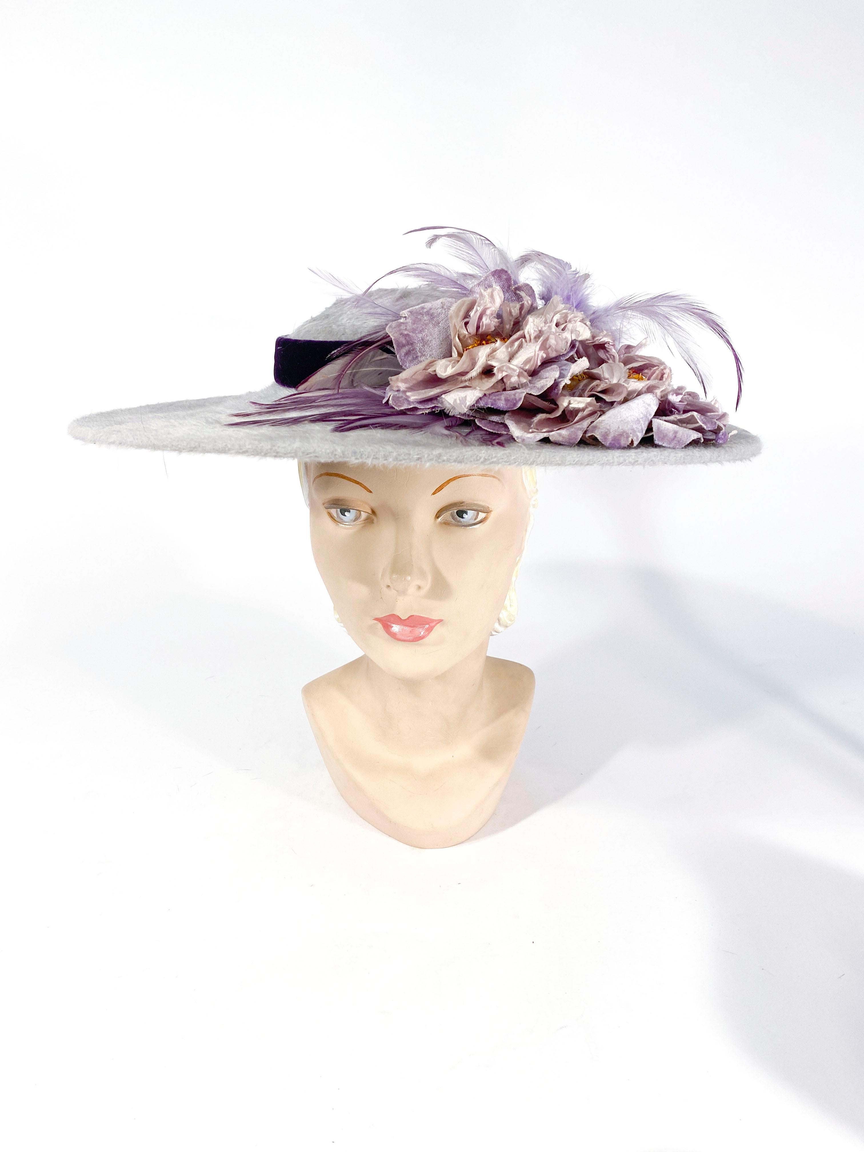1950s soft grey wide-brimmed picture hat made of beaver fur felt. The crown of the hat has a bold purple velvet band finished with handmade lavender silk velvet flowers and two-toned feathers. 