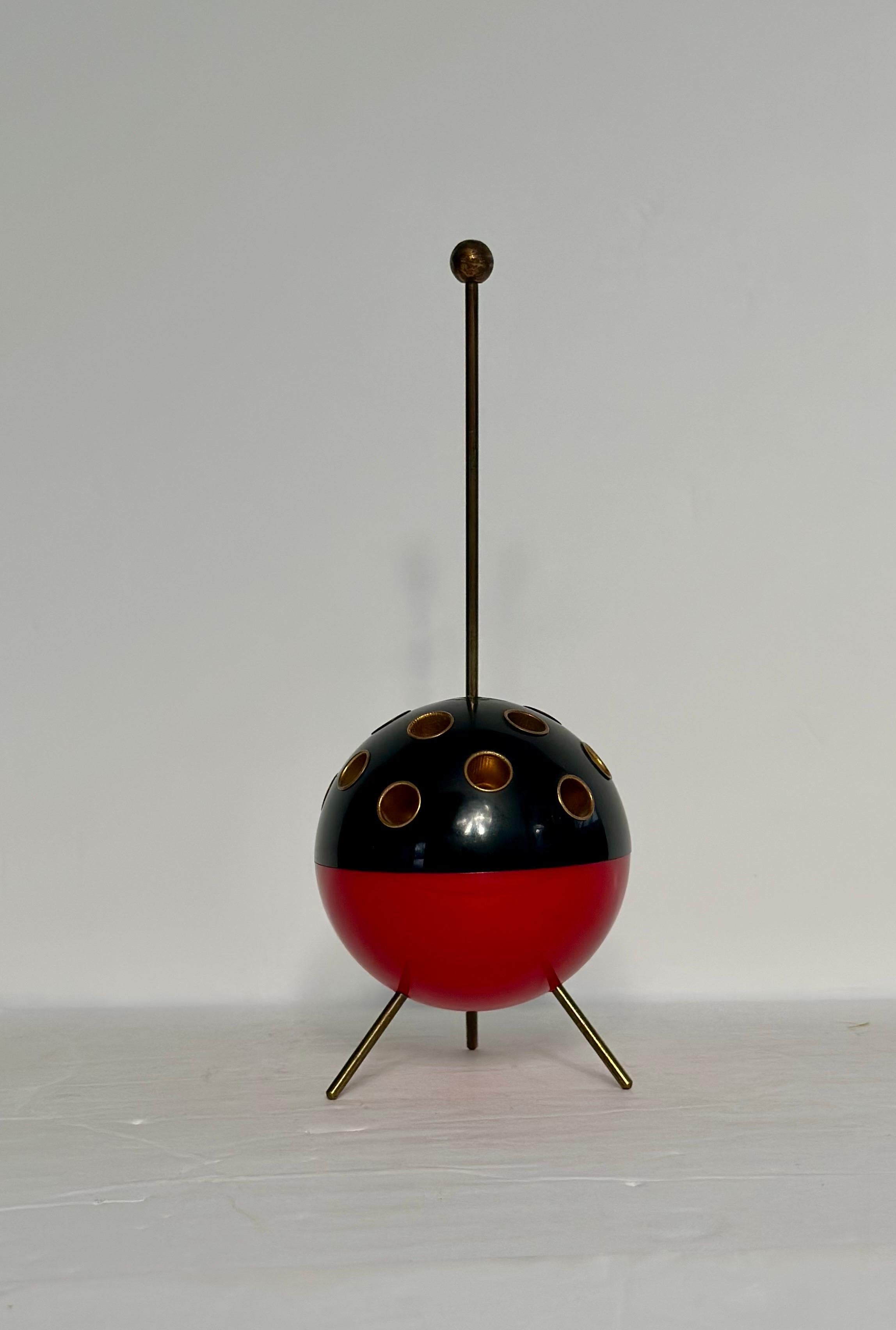 We are very pleased to offer a Mid-Century Modern Dublo cigarette dispenser circa the 1950s.  This stylish piece features a bold half-black, half-red ball with fifteen gold-plated inserts, sitting gracefully atop tripod brass legs that echo the