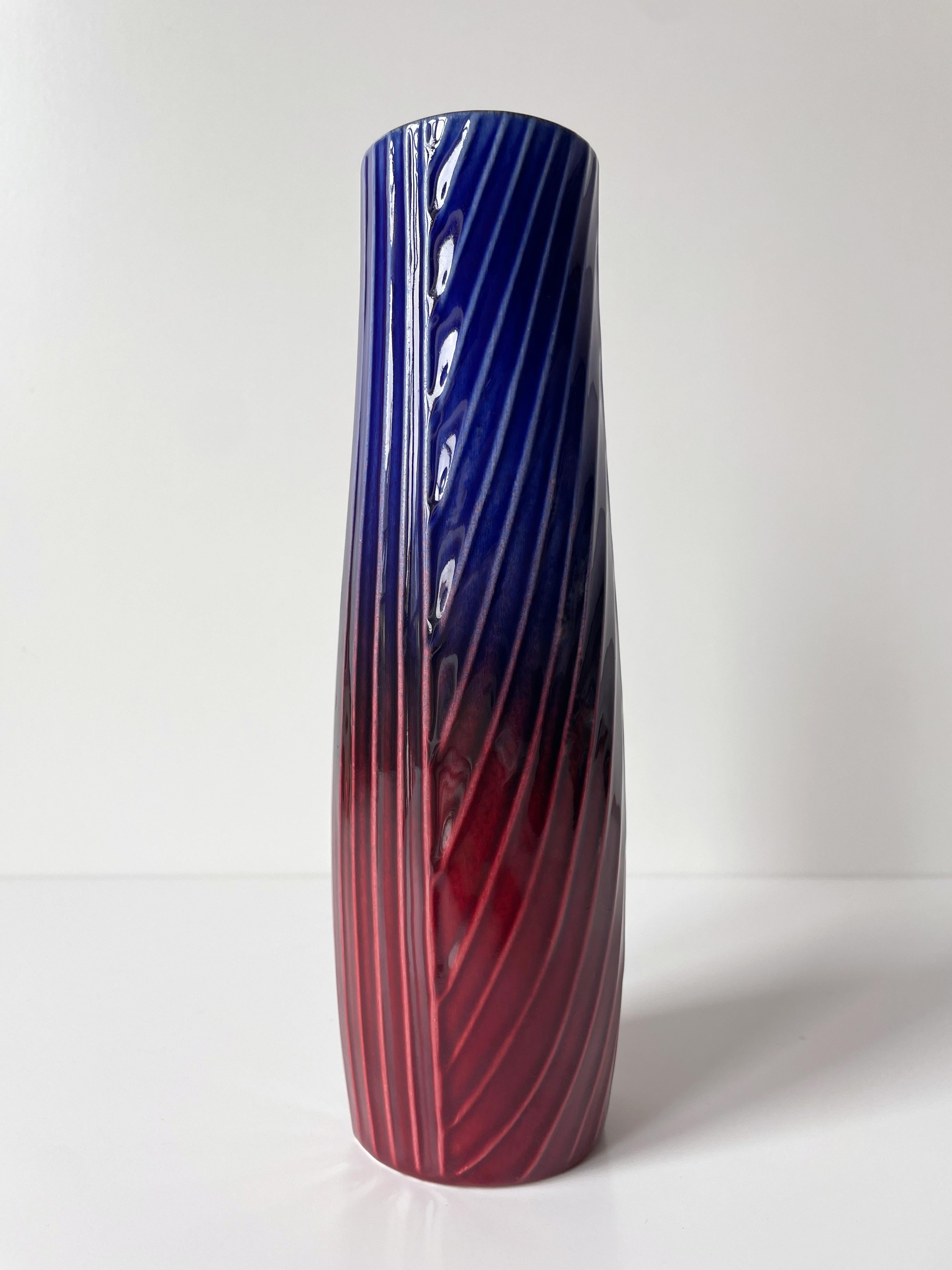 Slender cylinder shaped stoneware vase with glossy glaze in cobalt blue, deep purple and burgundy red over linear relief patterns. Acclaimed artist Carl-Harry Stålhane (1920-1990) designed the Sparaxis series - sometimes referred to as Anemone - for