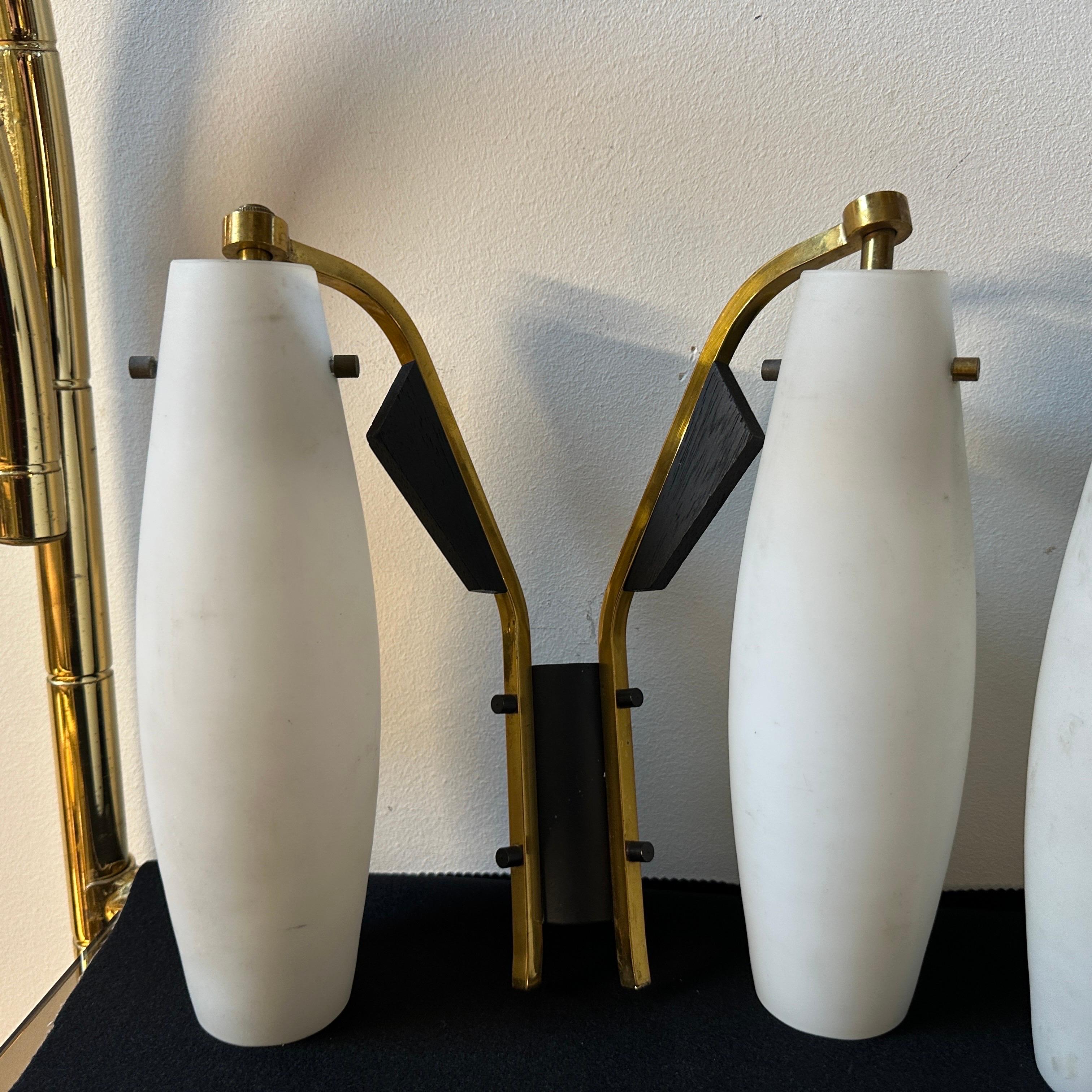 Two brass, black painted wood and white glass wall sconces designed and manufactured in Italy in the style of Stilnovo, they are in original patina and in working order. Crafted with brass, black painted wood, and metal, these sconces exhibit a