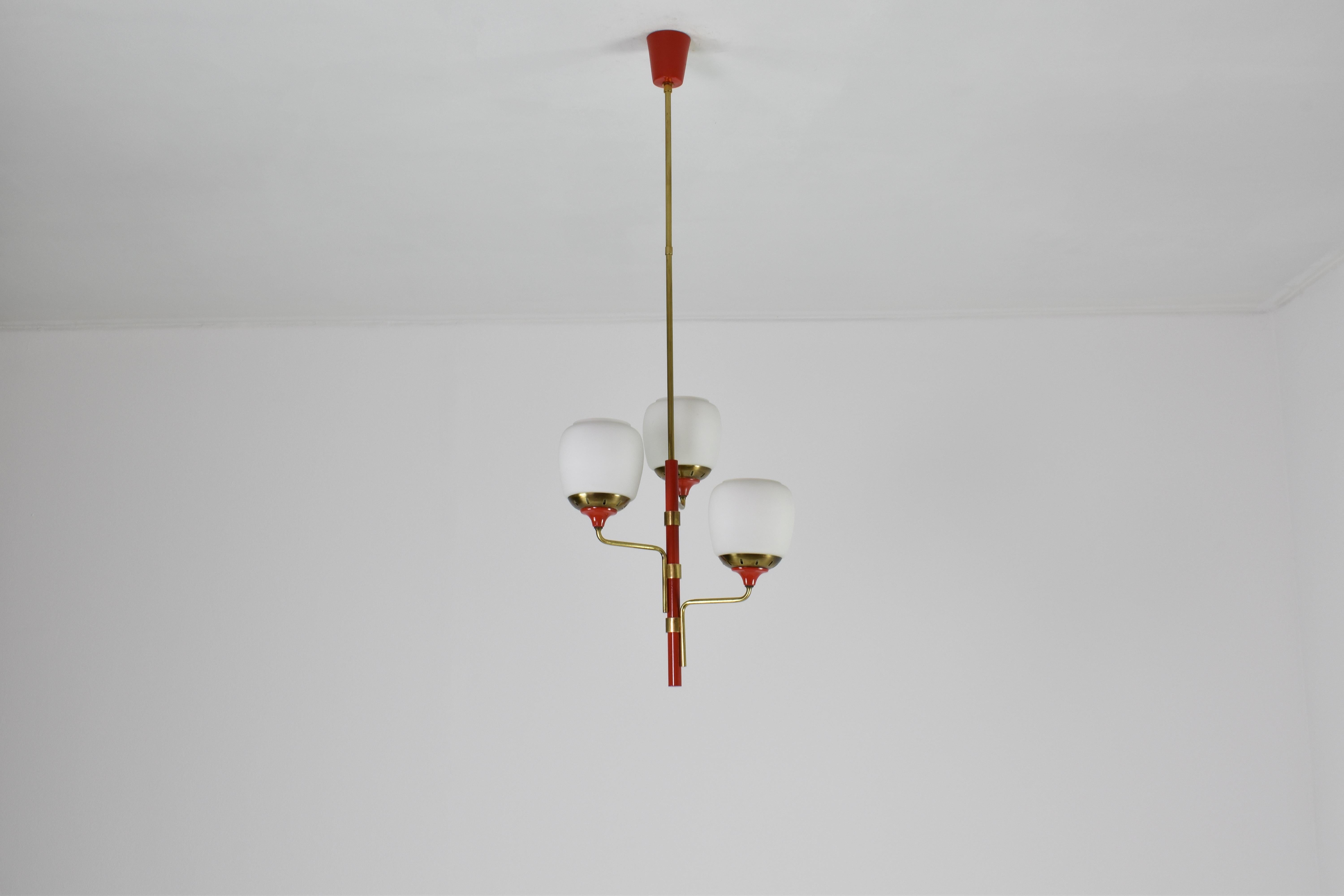 Cette suspension italienne des années 1950, attribuée à Stilnovo, se compose d'une tige en métal rouge laqué et de trois bras en laiton décalés, chacun supportant un globe en verre opalin et des colliers en laiton perforé.

La laque rouge injecte