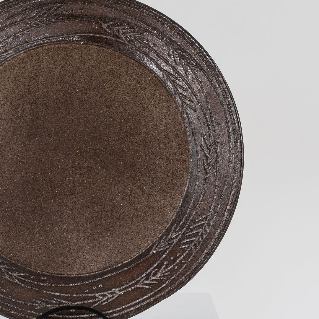 Il s'agit d'une grande assiette en grès, exécutée par Nancy Wickham Boyd pour son atelier du Vermont dans les années 1950. L'assiette est formée de grès brun glacé au sel et présente un motif de flèche directionnelle incisé. La pièce est signée