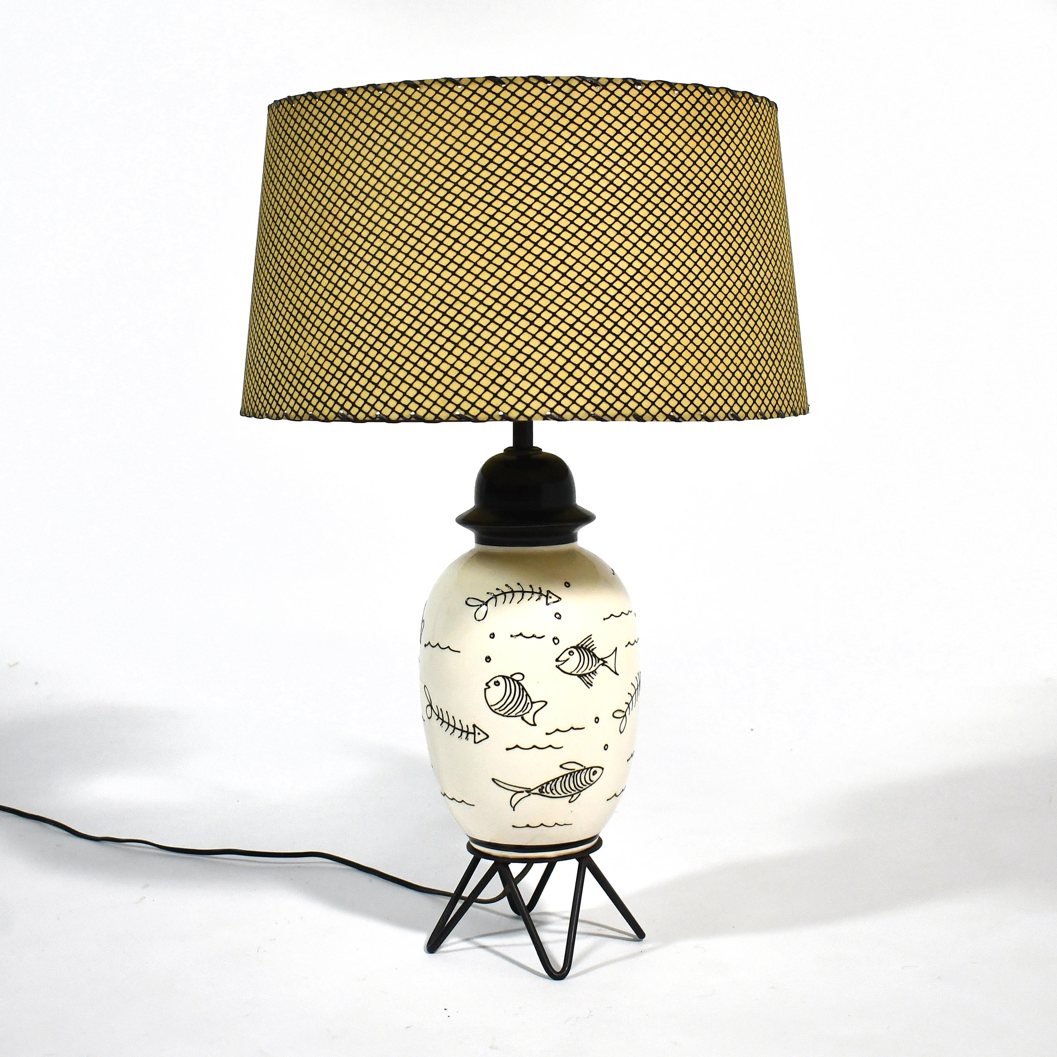 This playful table lamp has several qualities of 1950s design: the ceramic body has an urn-like form, is decorated with a whimsical fish design, and is supported by iron rod hair-pin legs. It is topped with a wonderful shade with a fishnet