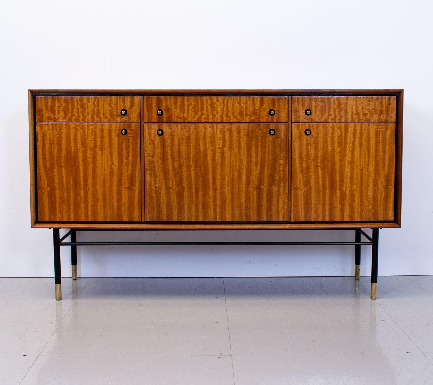 1950s tola and mahogany sideboard by Heals of London. It has 3 drawers, one baize lined for cutlery, 2 cupboards with removable shelves and a drinks cabinet with Formica lined door all on black painted steel legs with brass ferrules and handles.