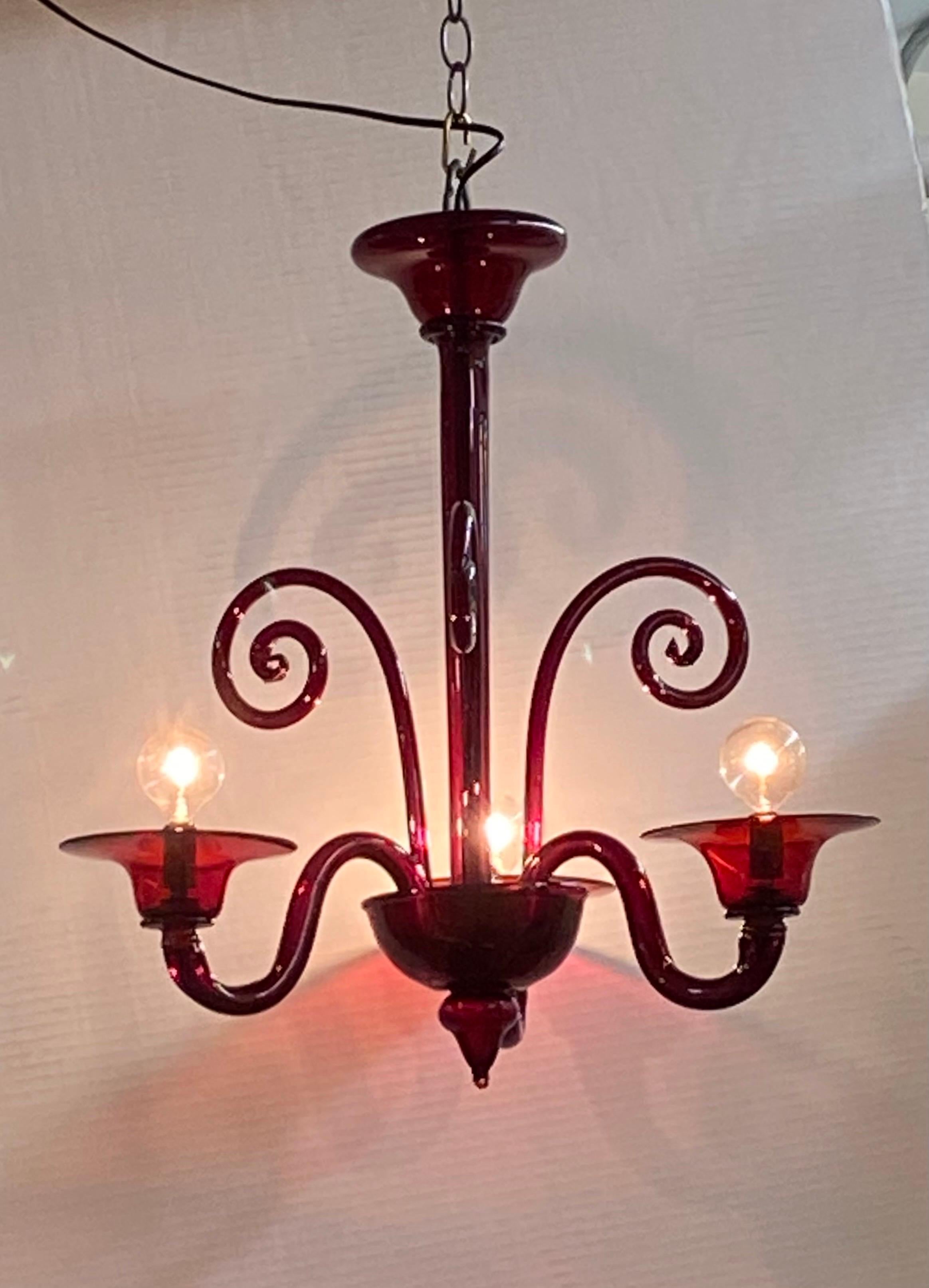A charming petite 1950s Venetian chandelier of hand blown glass in a stunning burgundy red color. The chandelier has three arms with each holding a candelabra socket alternating with three decorative tall rods ending in a spiral. The chandelier is