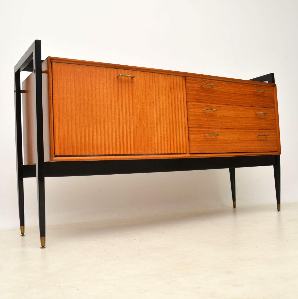 A stunning vintage sideboard, this dates from the 1950s-1960s. Its made from a light wood that looks like walnut, though it could be a light mahogany or tola wood. The legs are polished black with brass fixtures and this has brass handles as well.