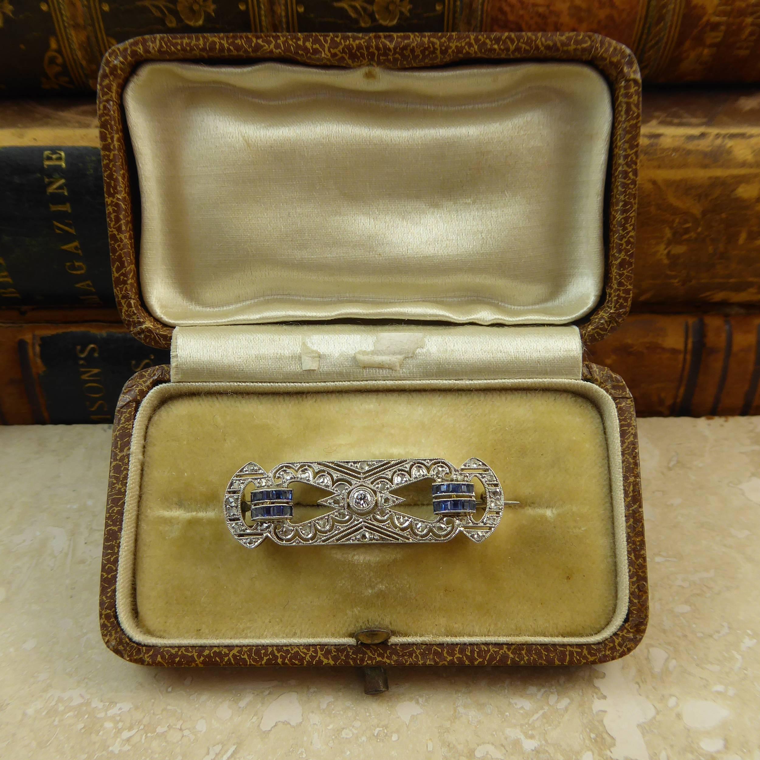 A mid-century sapphire and diamond brooch set to the centre with a round brilliant cut diamond approx. 2.0mm diameter to an open pierced diamond set surround of rougly rectangular shape.  A D-shaped, rose-cut diamond section to either end of the
