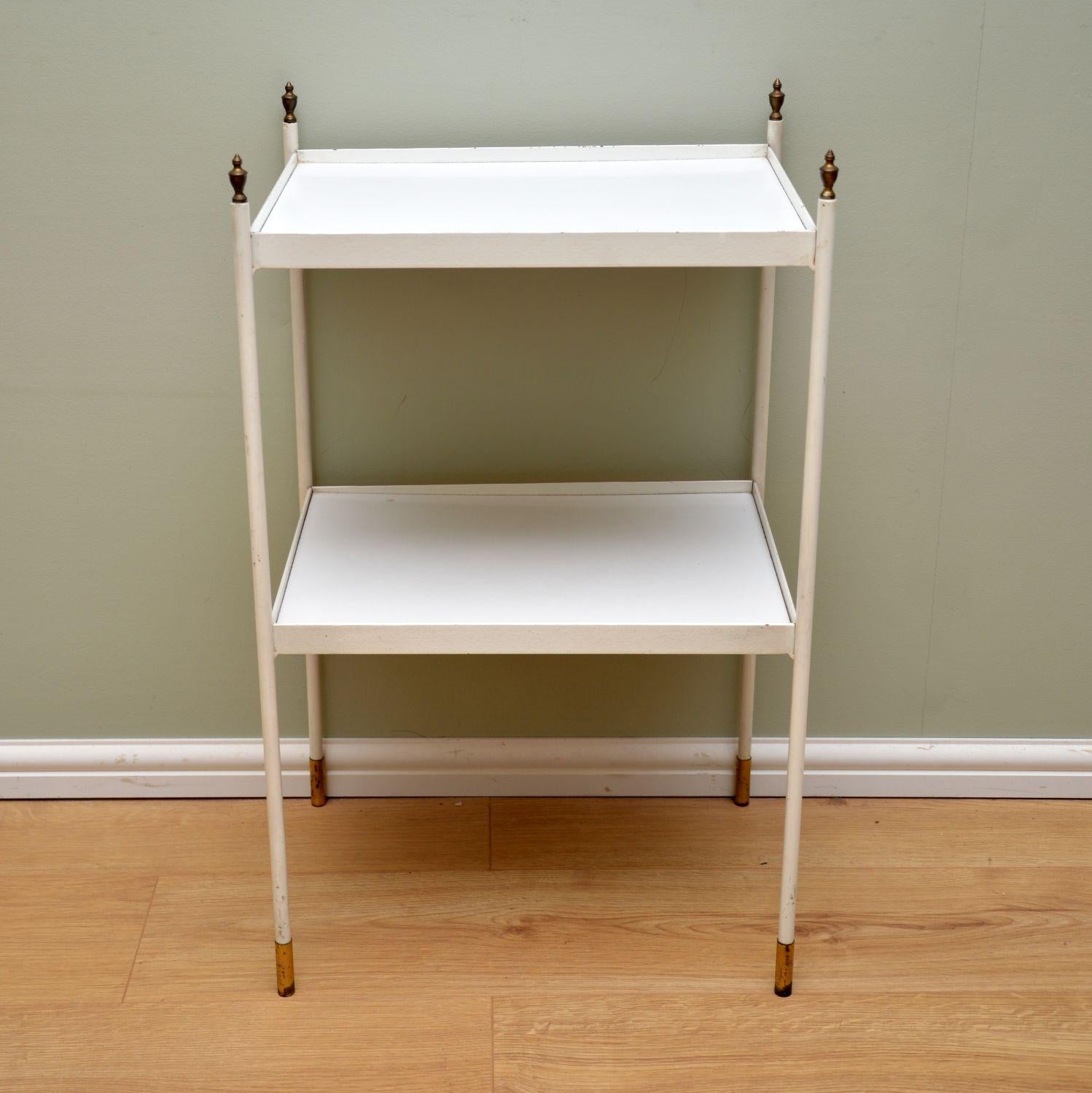 A stylish and elegant vintage two-tier side table. This dates from circa 1950s-1960s, it’s of lovely quality.

This has a painted white steel frame, with brass finials and feet caps. The shelves are wood with white Formica tops.

The condition