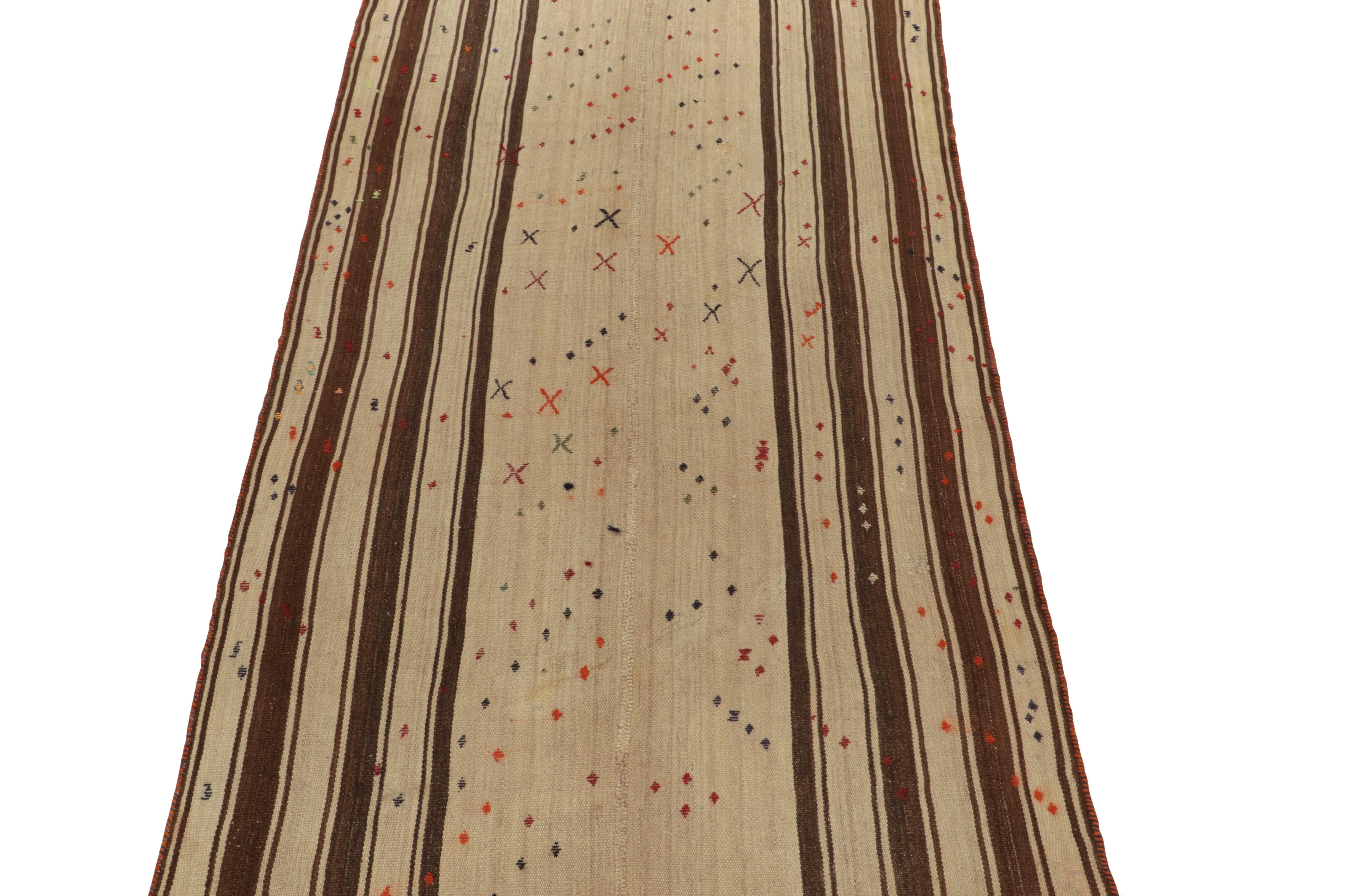 Originating from Turkey circa 1950-1960, a 5x10 mid-century kilim rug now entering our antique & vintage flat weaves curations. Between beige-brown stripes in vertical borders, the rug field enjoys colorful geometric patterns for a