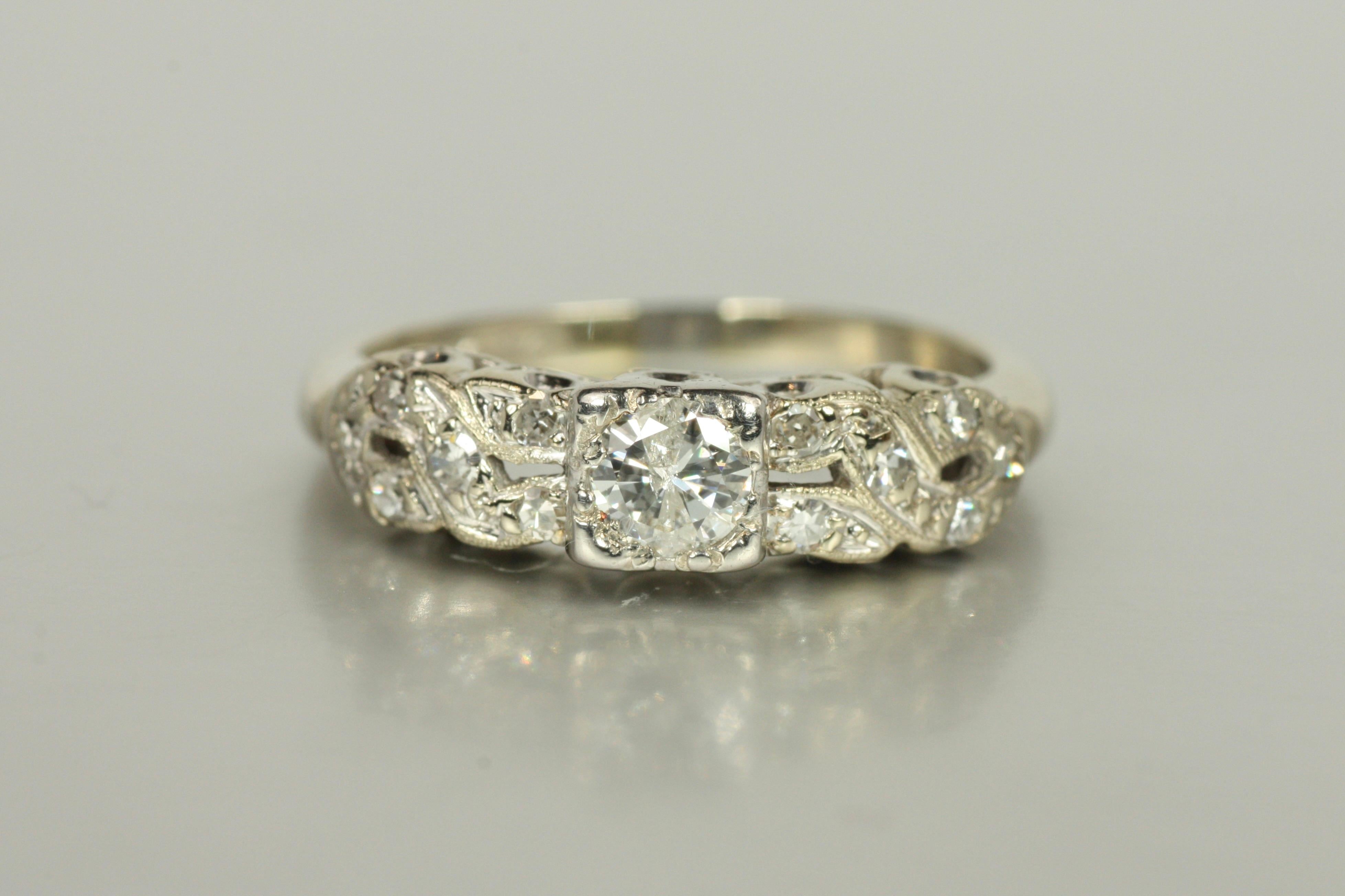 Un anello di fidanzamento vintage unico, con un diamante rotondo da 0,20 carati circondato da altri 12 diamanti che decorano un design intrecciato in oro bianco 14 carati. Realizzato negli anni '50, questo anello è un'ottima rappresentazione