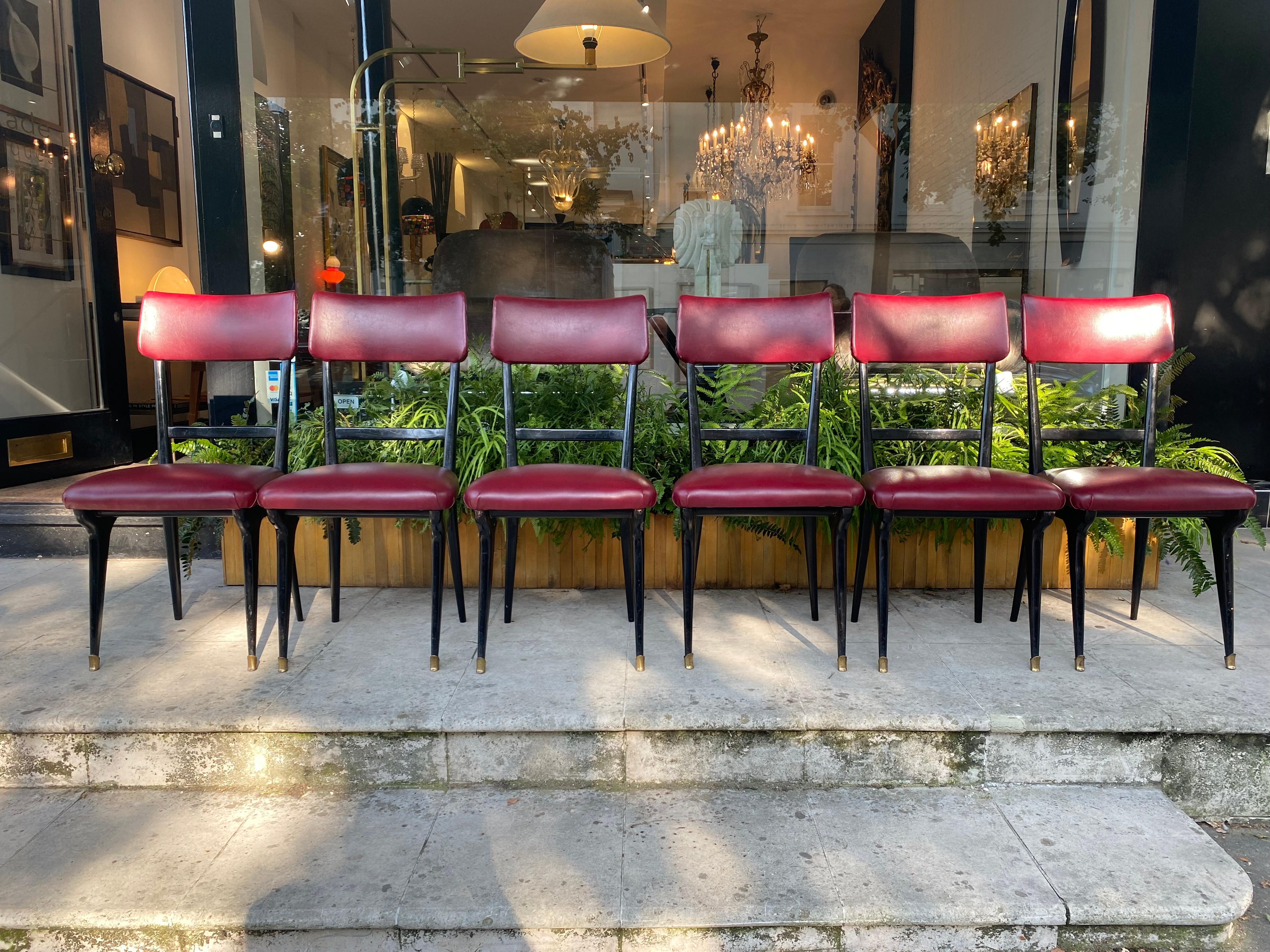 A set of 6 dining chairs by Vittorio Dassi upholstered in their original faux leather Dubonnet red on black lacquered frame with brass sabots. The chairs have got a subtle curve to the back rest and lovely curved details to to the top of the front