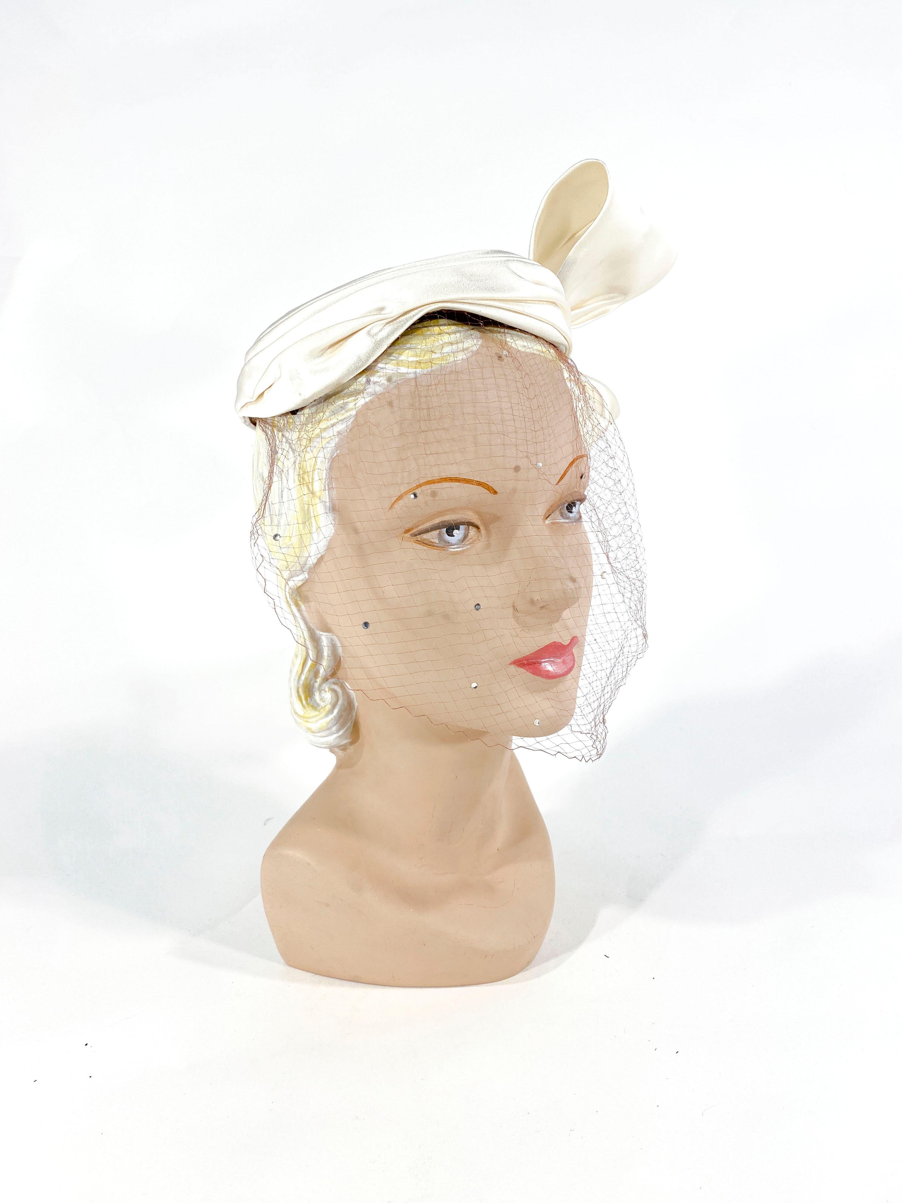 1950s White silk satin cocktail hat with an enlarged sculpted bow that finished the gathered edge at the base of the hat. The full-face veil is a soft tan color decorated with clear rhinestone accents.