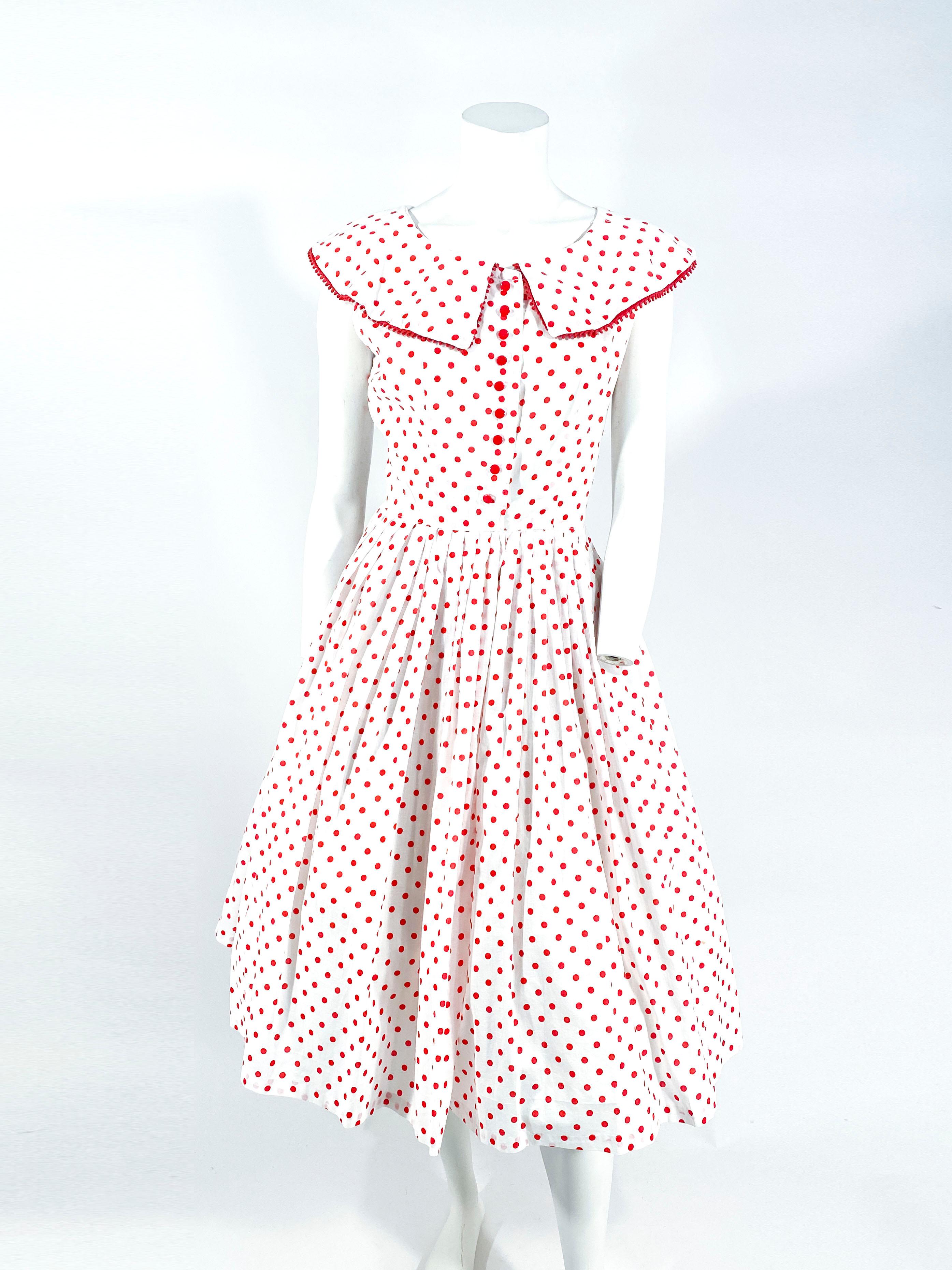 1950s white cotton day dress featuring a red polka dot print, puritan collar, bric-a-brac trim, front button closure, plastic buttons, and a matching plastic belt with cut-outs. The skirt is very full and has several knife pleats along the waist