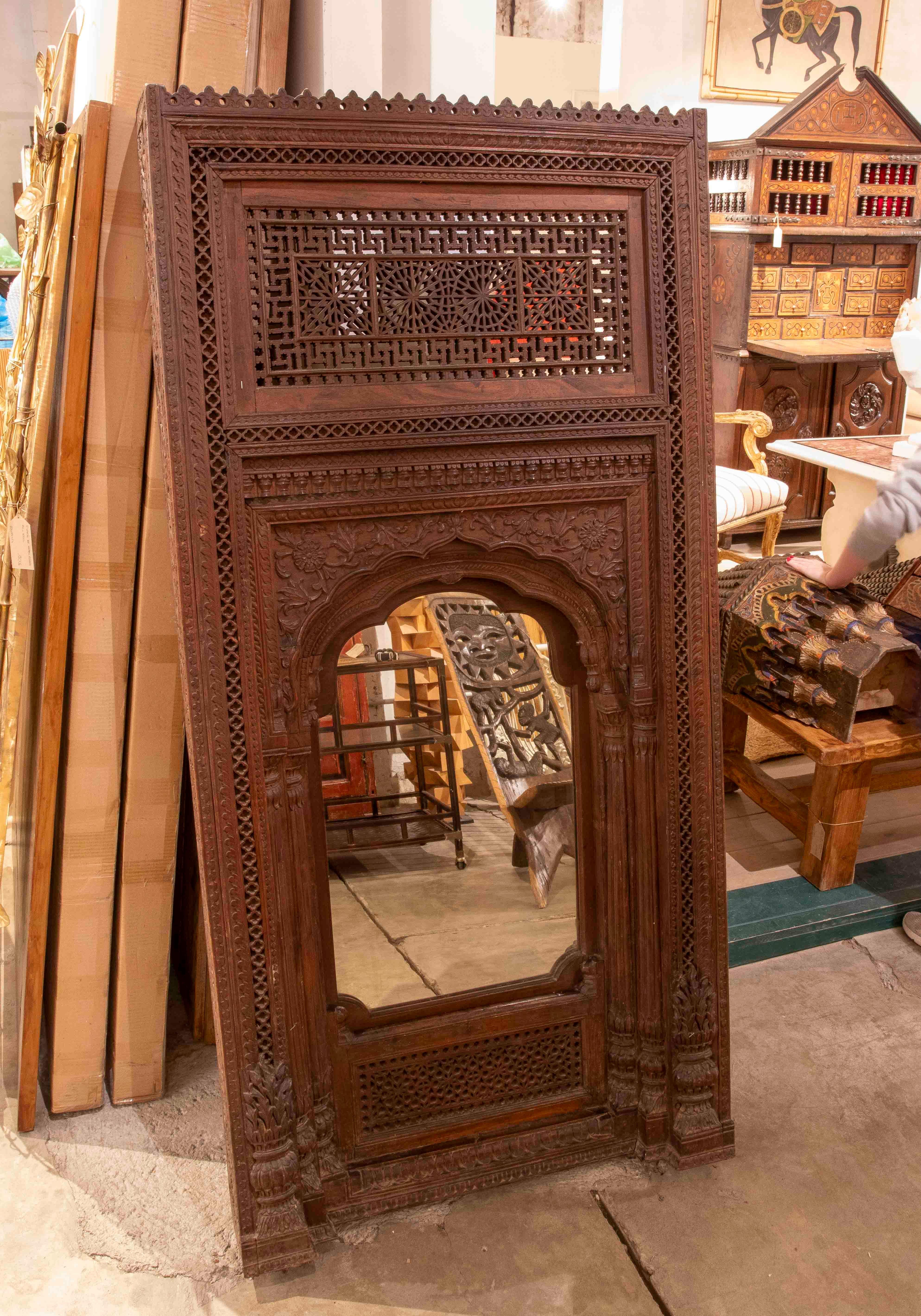 Superbe miroir ancien avec un cadre architectural en bois massif sculpté à la main, inspiré de l'architecture INDO-. Son design complexe présente des motifs géométriques et floraux, des arcs lobés et des détails ajourés qui témoignent d'un