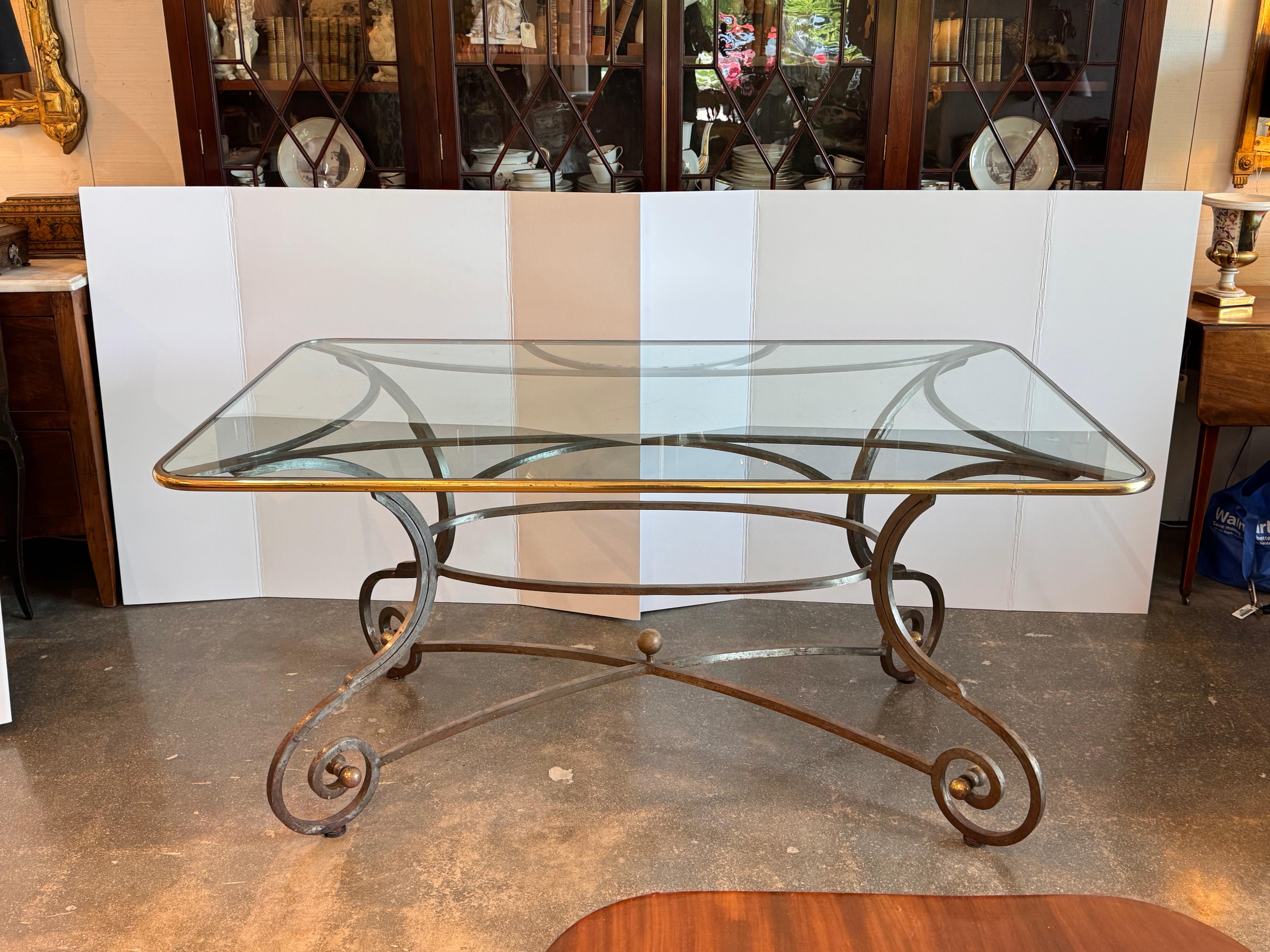 Rectangular glass-top table with a scrolled steel base. The clear glass is framed by a subtle gilt edge, resting atop an elegant iron structure with decorative curled feet and central orb details. The curvilinear form evokes traditional European