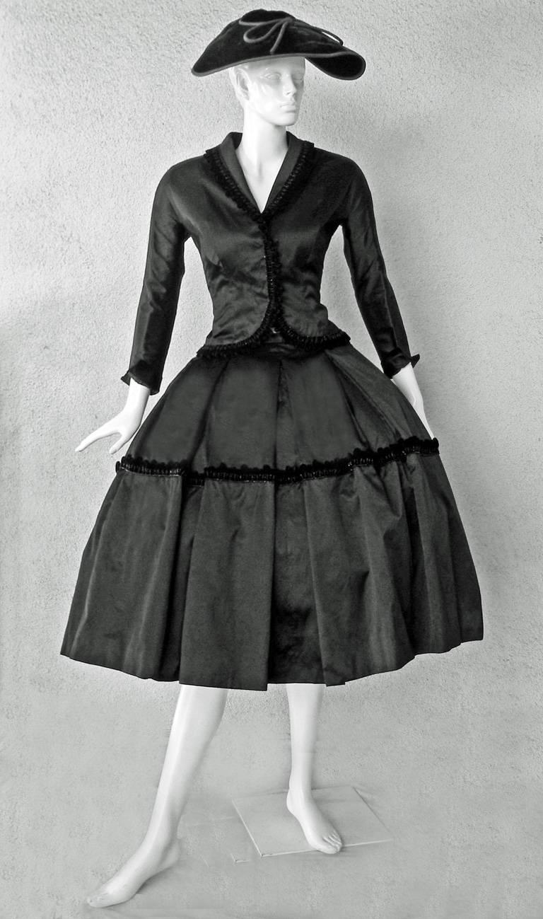 Two piece suit consisting of a jacket and mid length skirt of butter black satin trimmed with a velvet and jet fringe. The fitted jacket is constructed on princess lines with raglan sleeves, a cutaway closure and small peplum. The full ballerina